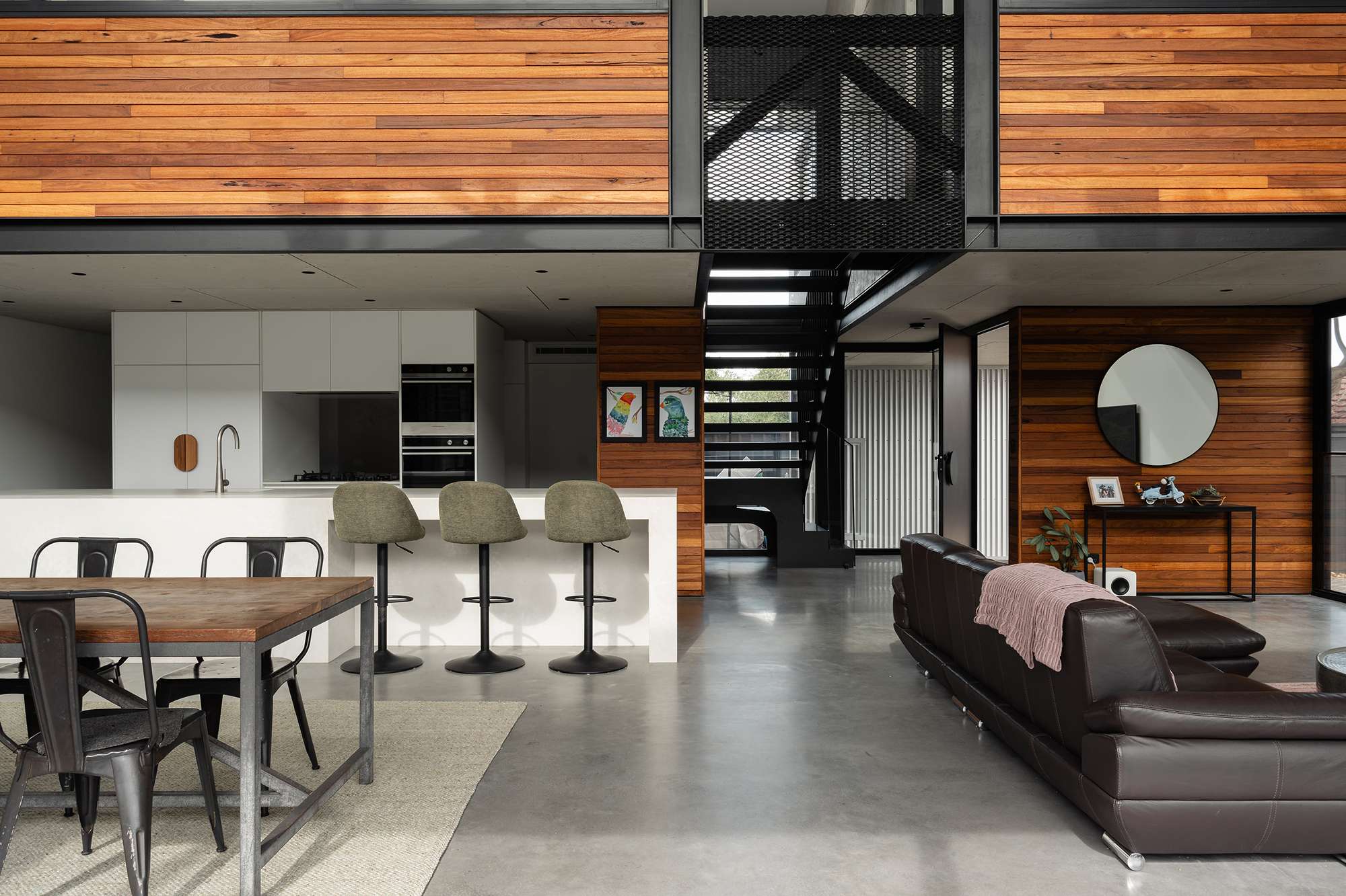 Open-plan kitchen, dining and living space with polished concrete flooring, timber wall cladding and a black steel staircase leading to an upper level.