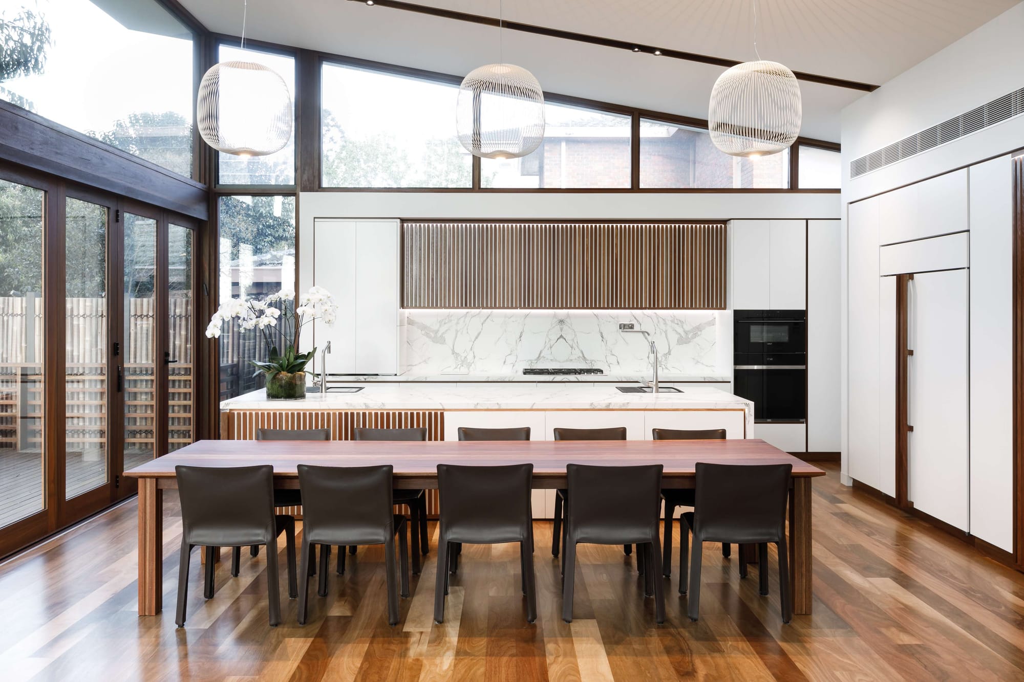Kitchen and dining space with timber flooring, battened joinery and a long dining table beneath a sloping ceiling, with tall glazing framing the garden.