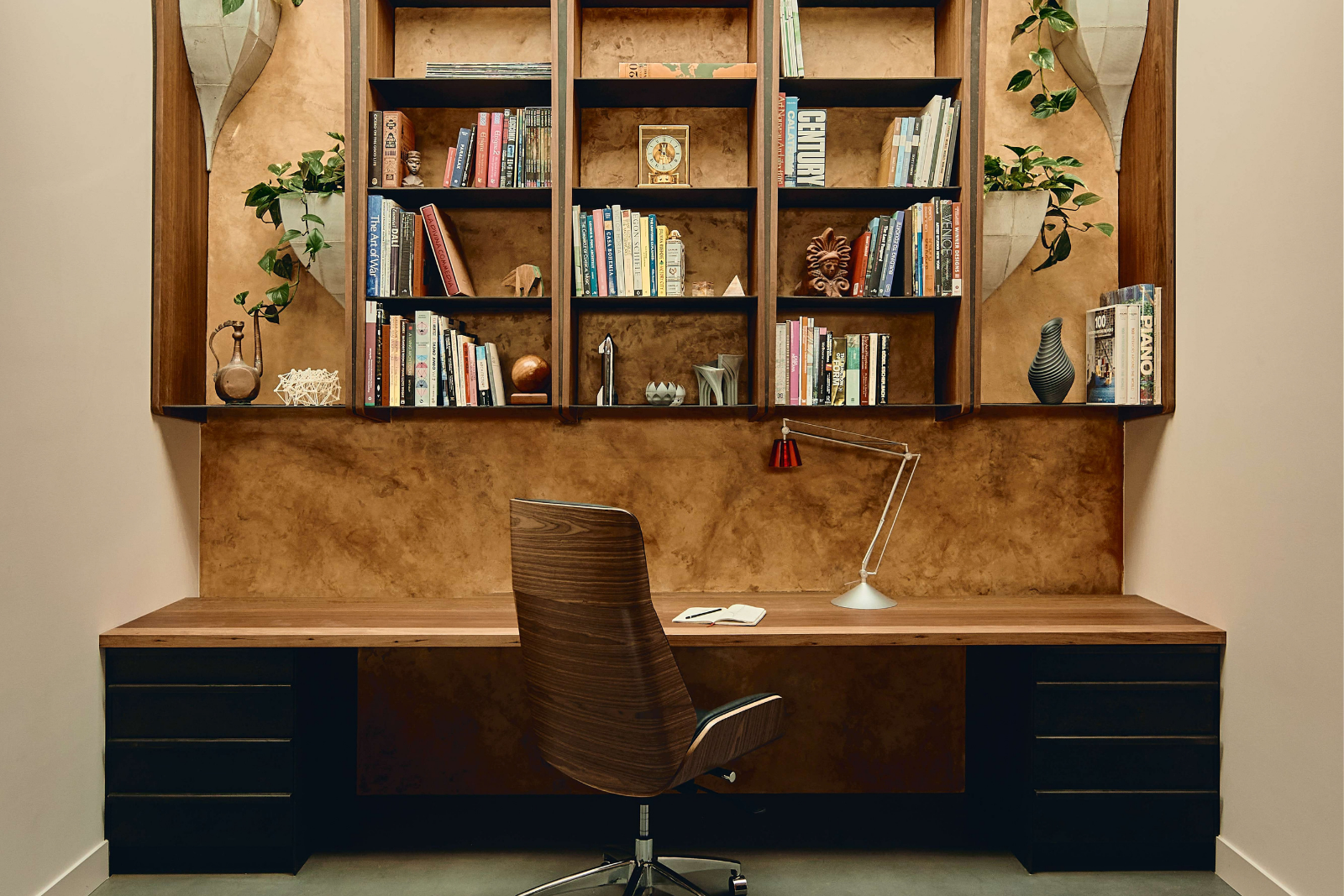 Built-in timber desk with dark drawer units, a timber swivel chair and open bookshelves against an ochre wall, framed by hanging planters.