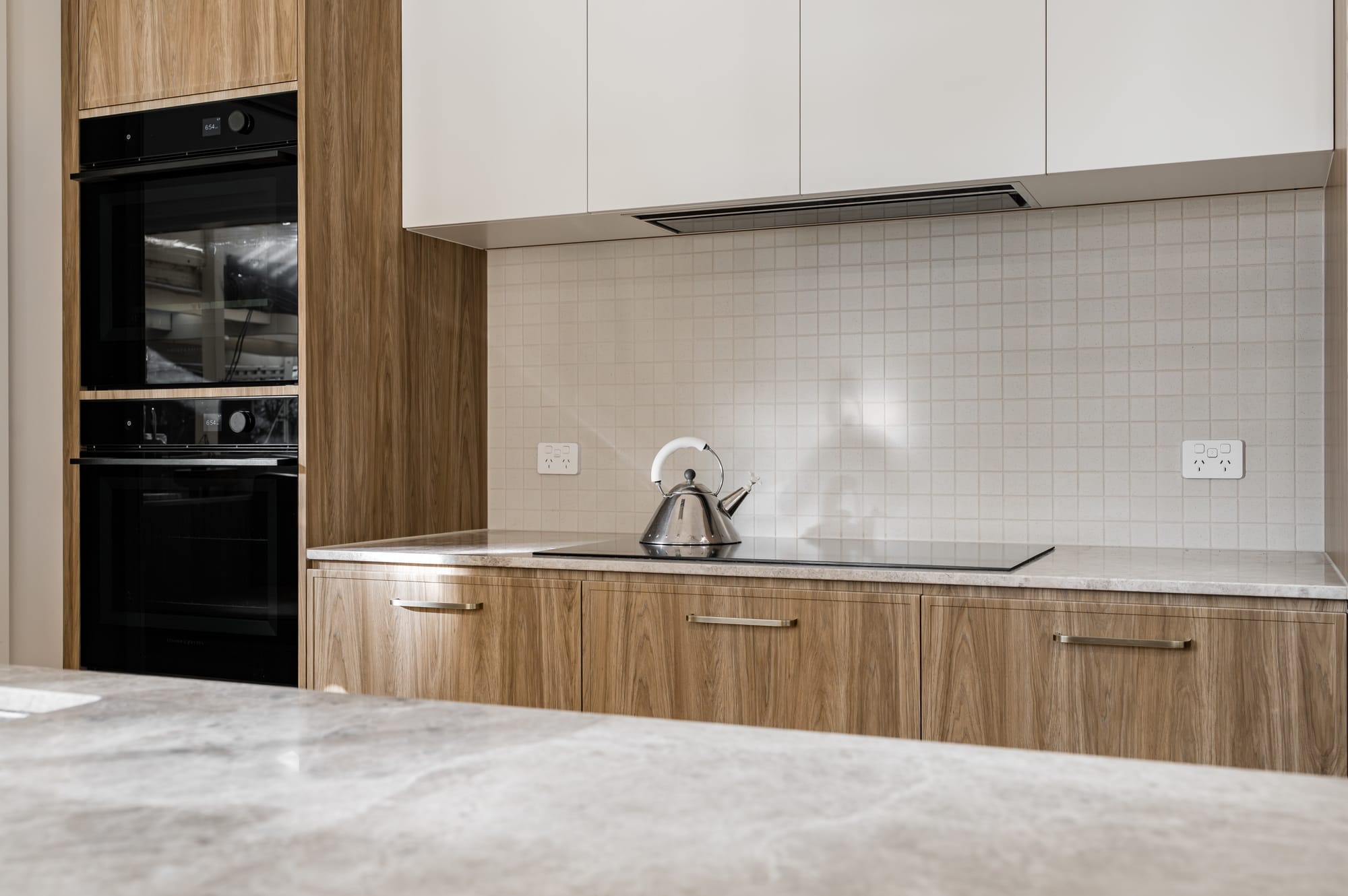 Kitchen with timber look cabinetry, stacked black ovens, a pale benchtop and white square tile splashback, lit by natural light.