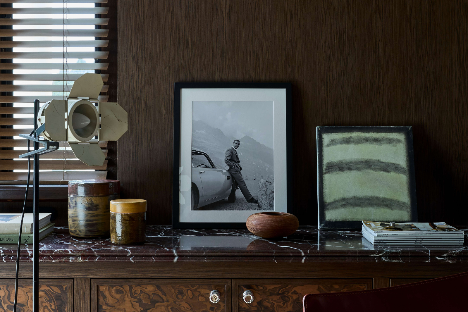 Dark timber cabinetry with a veined stone benchtop displaying framed artworks, ceramic vessels and stacked magazines beside a tripod spotlight and Venetian blinds.
