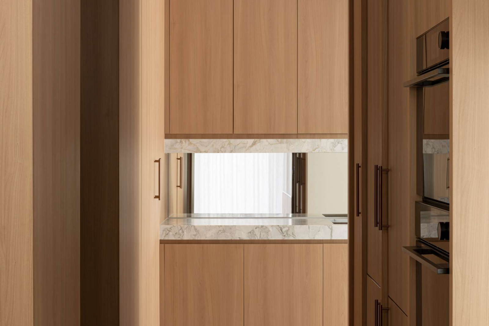 View through a doorway into a kitchen lined with timber cabinetry, featuring long bronze toned handles, integrated ovens and a pale stone benchtop.