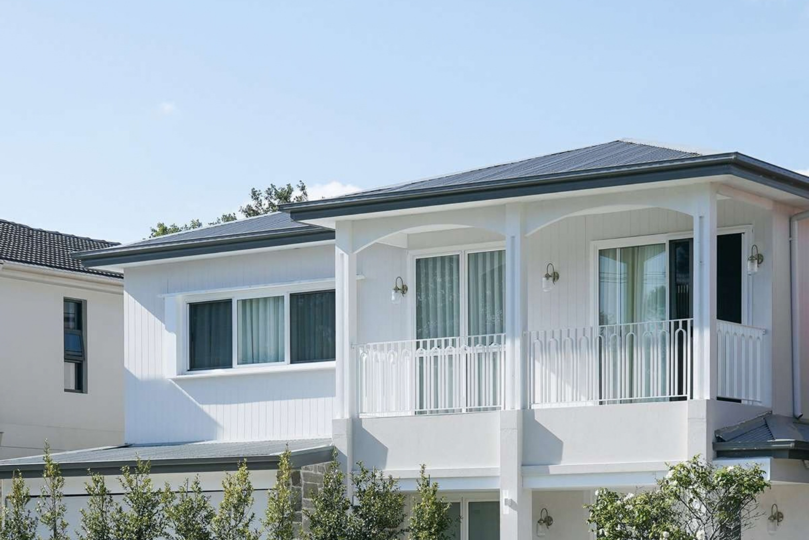 White two-storey house with vertical cladding, a dark metal roof and a covered upper balcony with decorative white balustrades.