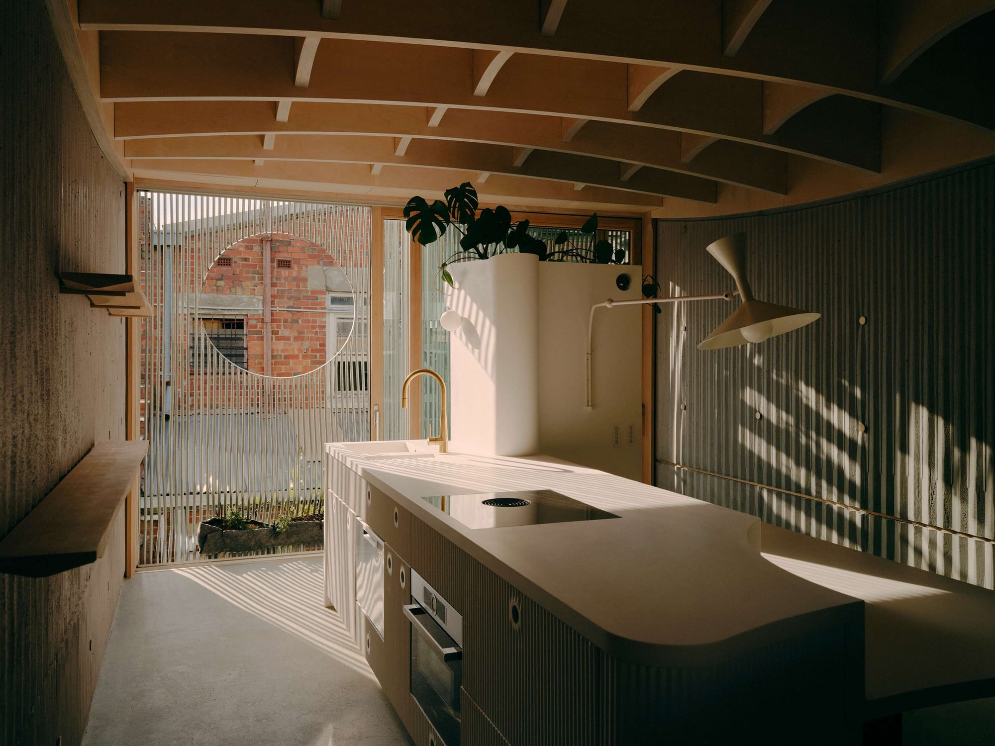 Sunlight filters through a vertical screen, casting striped shadows across a pale kitchen island and textured concrete walls beneath a curved timber ceiling.