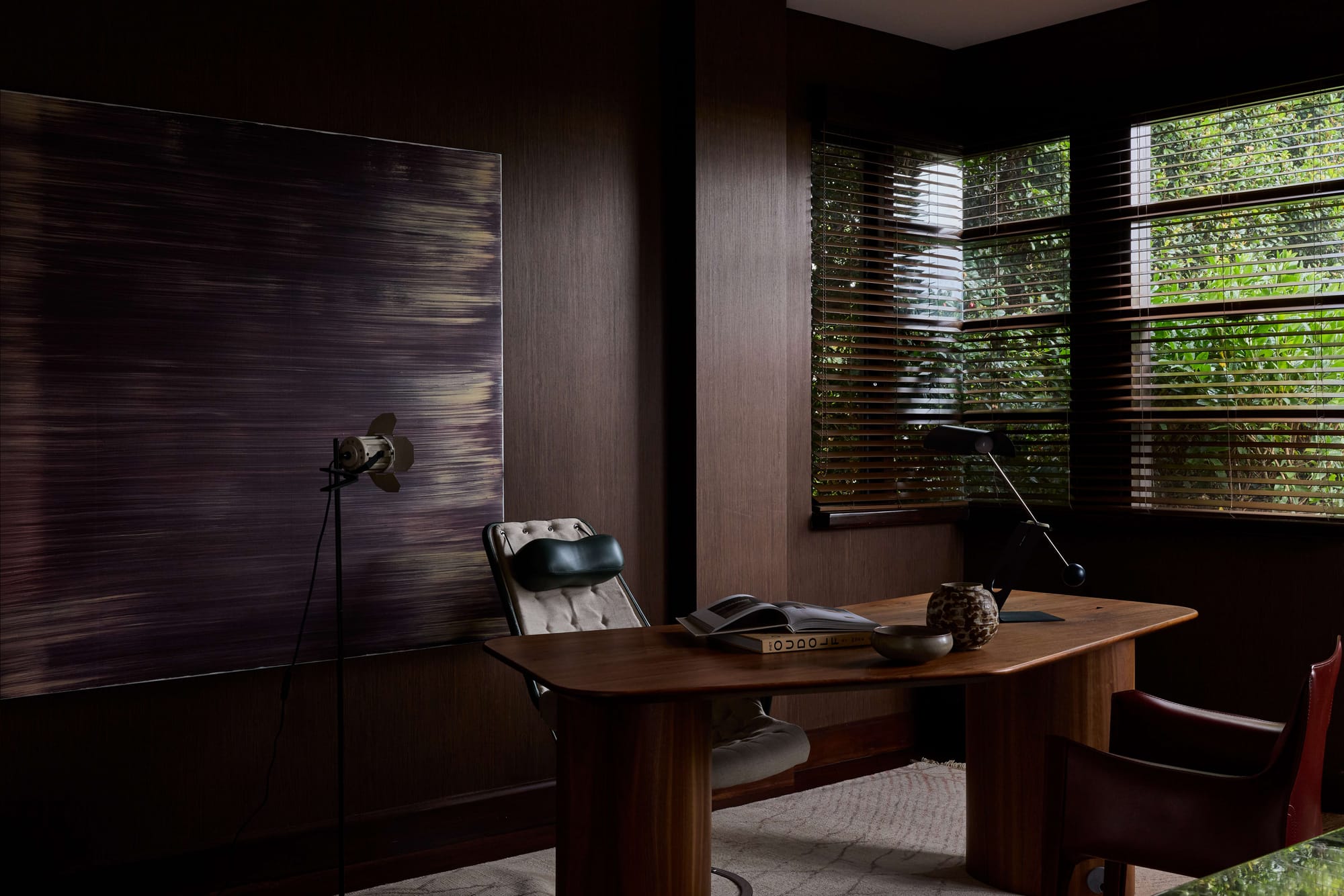 Home office at Rose Bay Residence by Studio PULP, with dark timber wall panelling, a curved timber desk, abstract artwork and corner windows with Venetian blinds overlooking greenery.