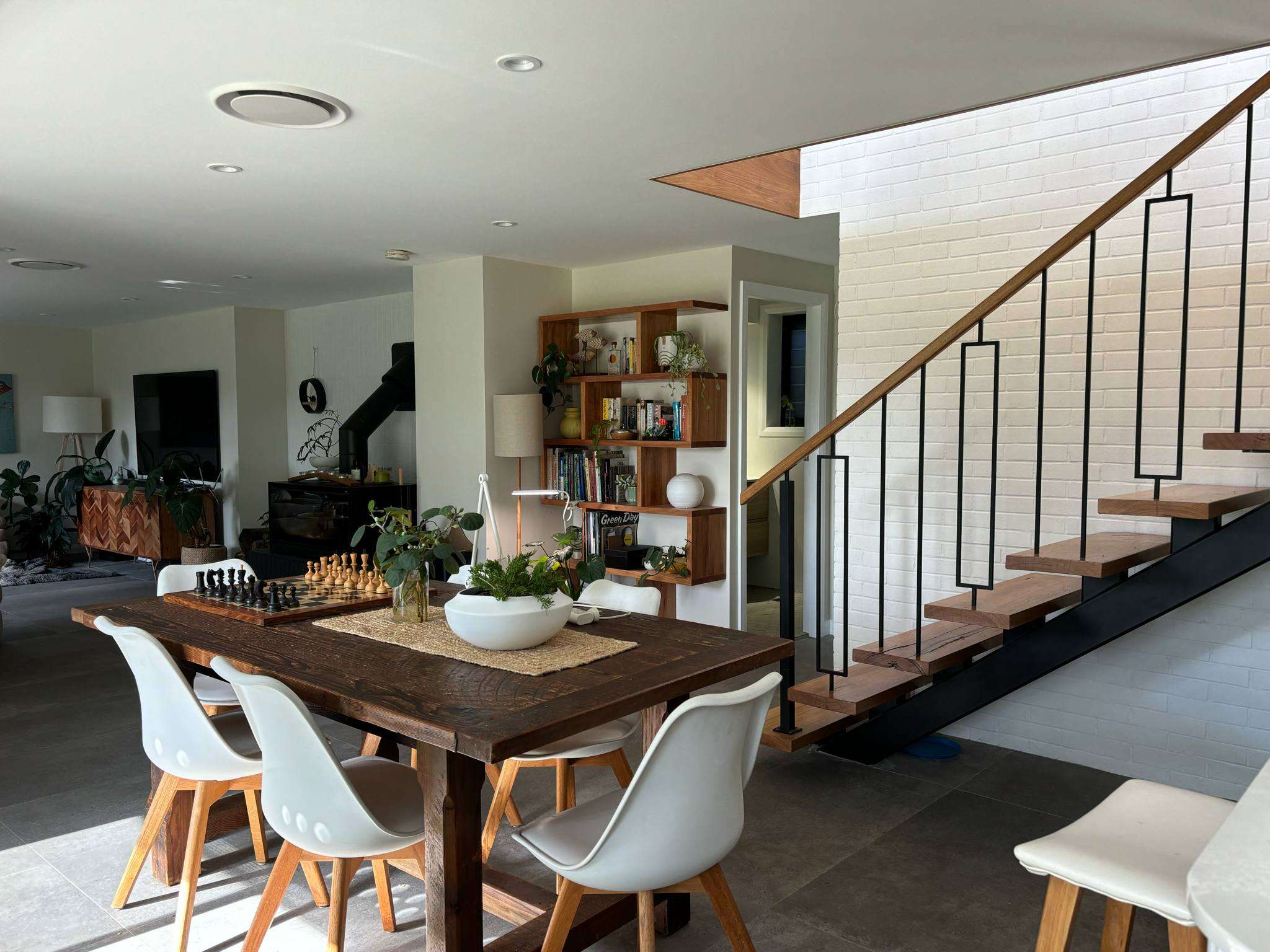 Sunlit dining area with a timber table, white chairs and a chessboard beside an open timber staircase with geometric black balustrades and a white brick wall.
