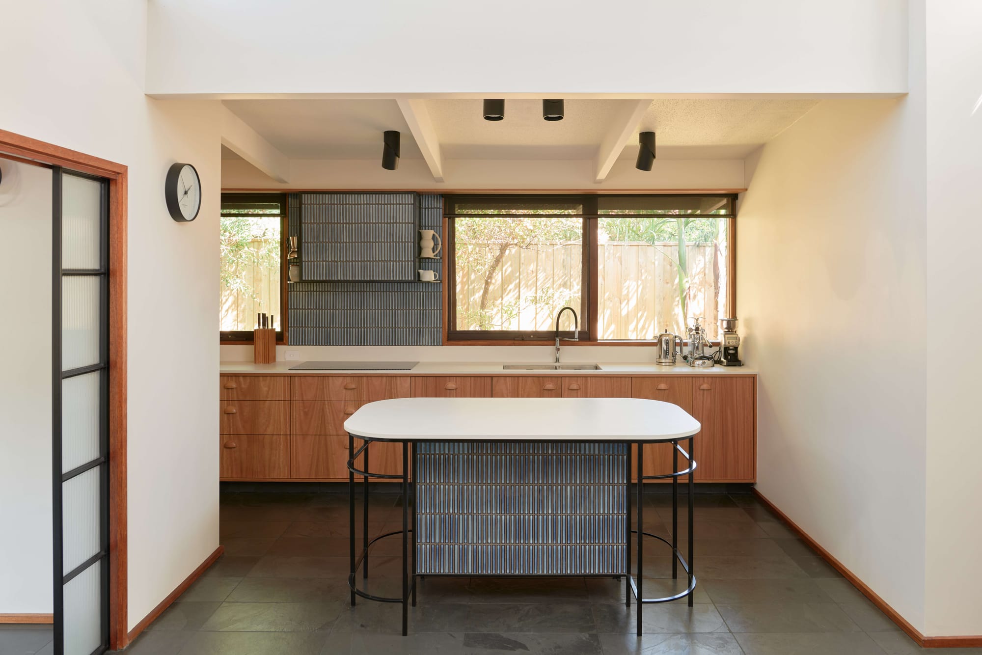 Kitchen with timber cabinetry beneath garden facing windows, dark tiled flooring and a rounded island with a pale top and slender black framework.