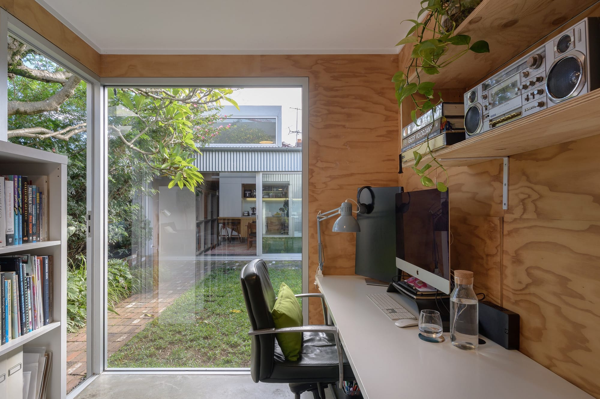 Plywood-lined home office with a white desk, computer and open shelving beside full-height corner glazing overlooking a leafy courtyard.