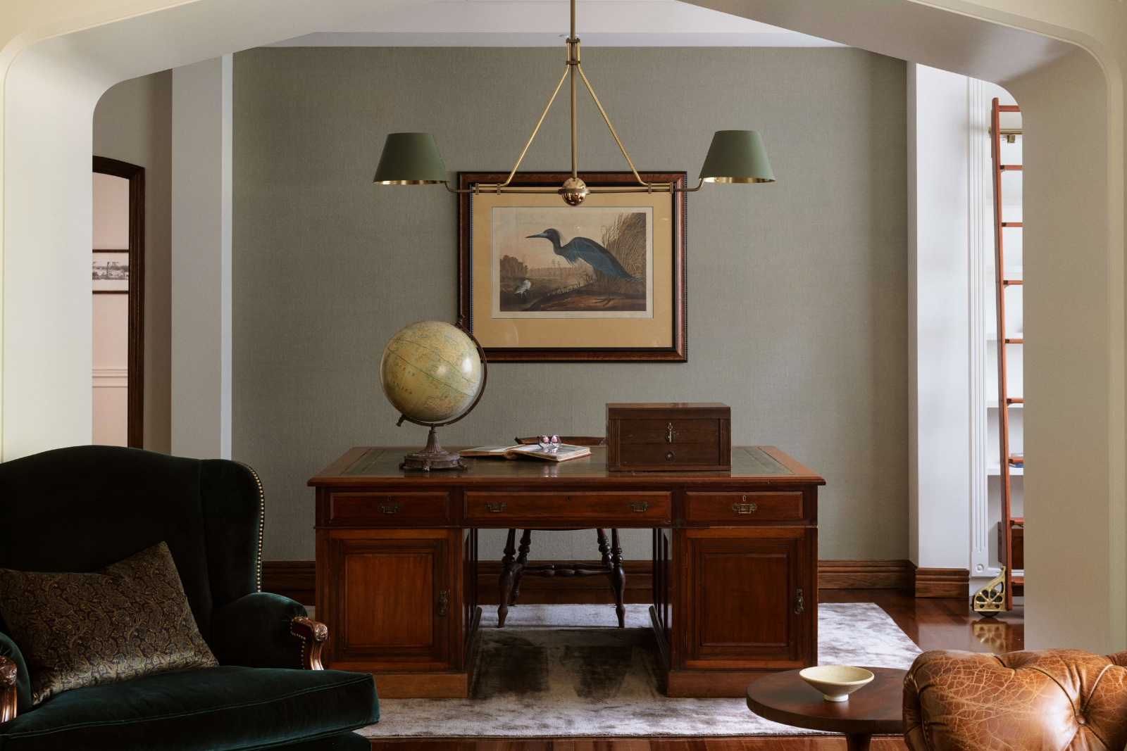 Traditional study framed by a broad curved opening, with a timber pedestal desk, globe, framed bird artwork and brass pendant with green shades.