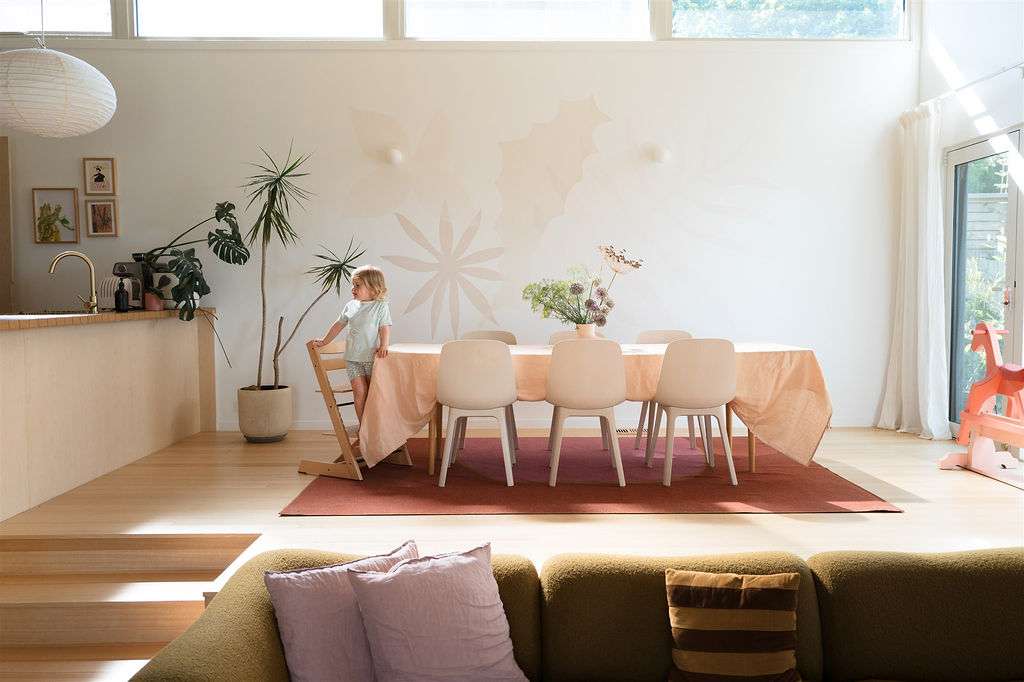 Sunlit dining area with pale timber joinery and flooring, a peach tablecloth and high windows, positioned above a sunken living space.