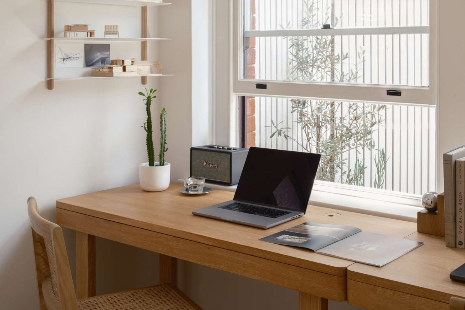 Pale timber desk beneath a large sash window, with a laptop, woven chair, potted cactus and wall shelves displaying architectural models.