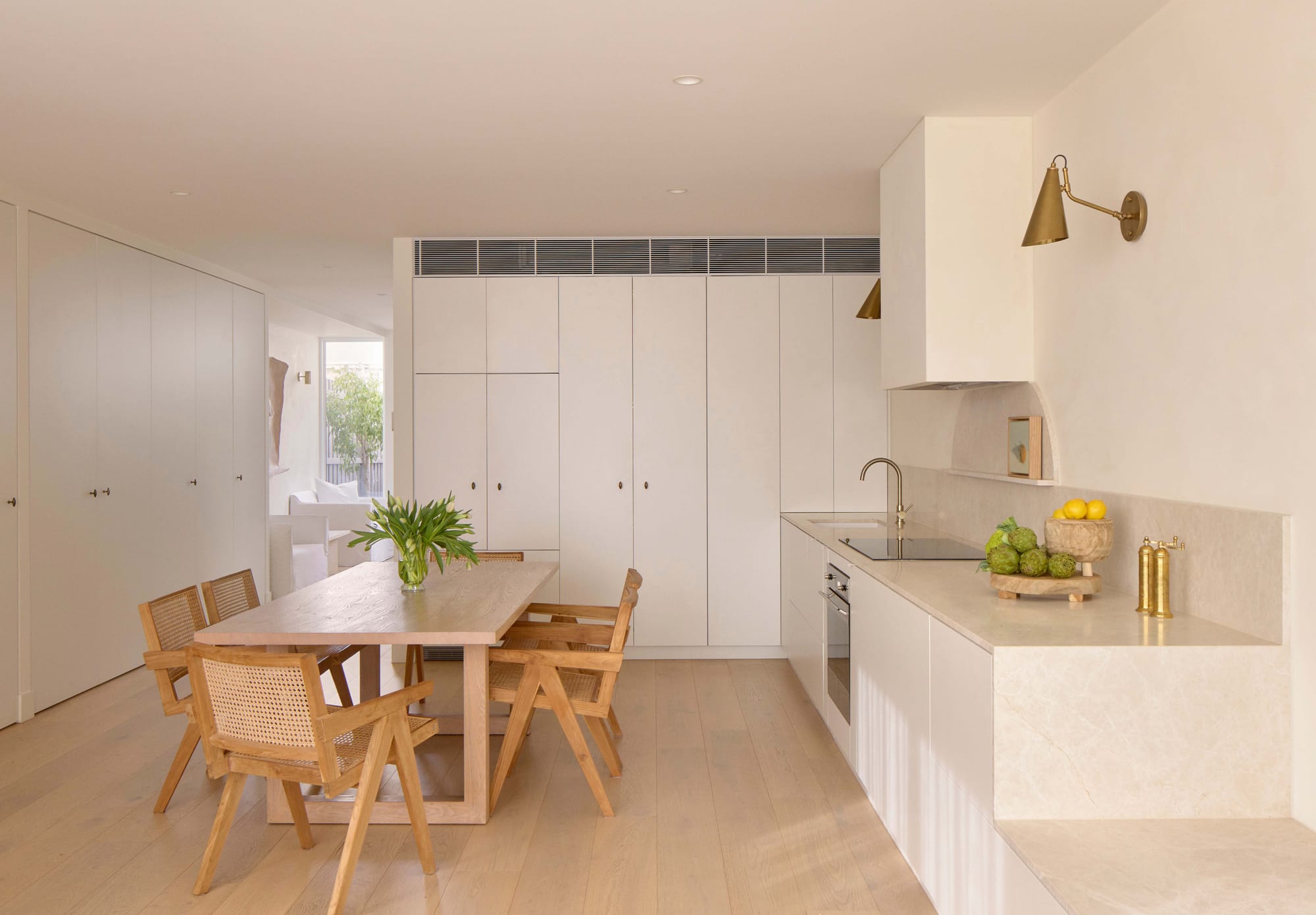 Kitchen and dining area with white cabinetry, a pale stone benchtop, brass wall lights and a timber dining table with woven chairs.