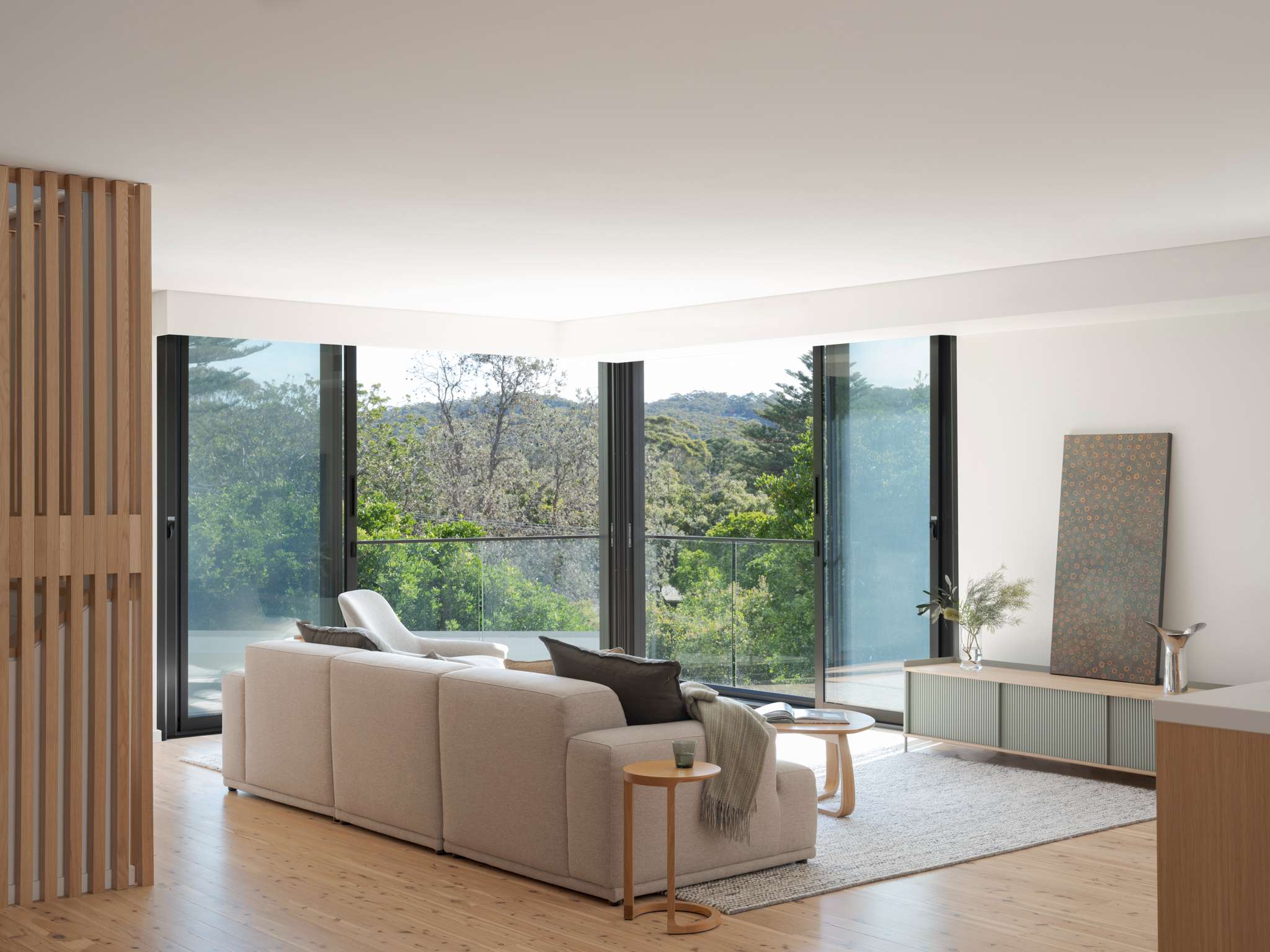 Light-filled living room with a pale modular sofa, timber flooring and full-height glazing overlooking a leafy landscape.