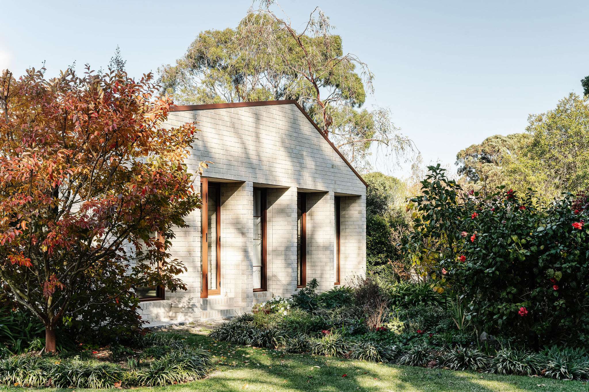 A pale brick addition with an angled roofline and tall, recessed windows sits within a leafy garden. Sunlight and tree shadows fall across the façade.
