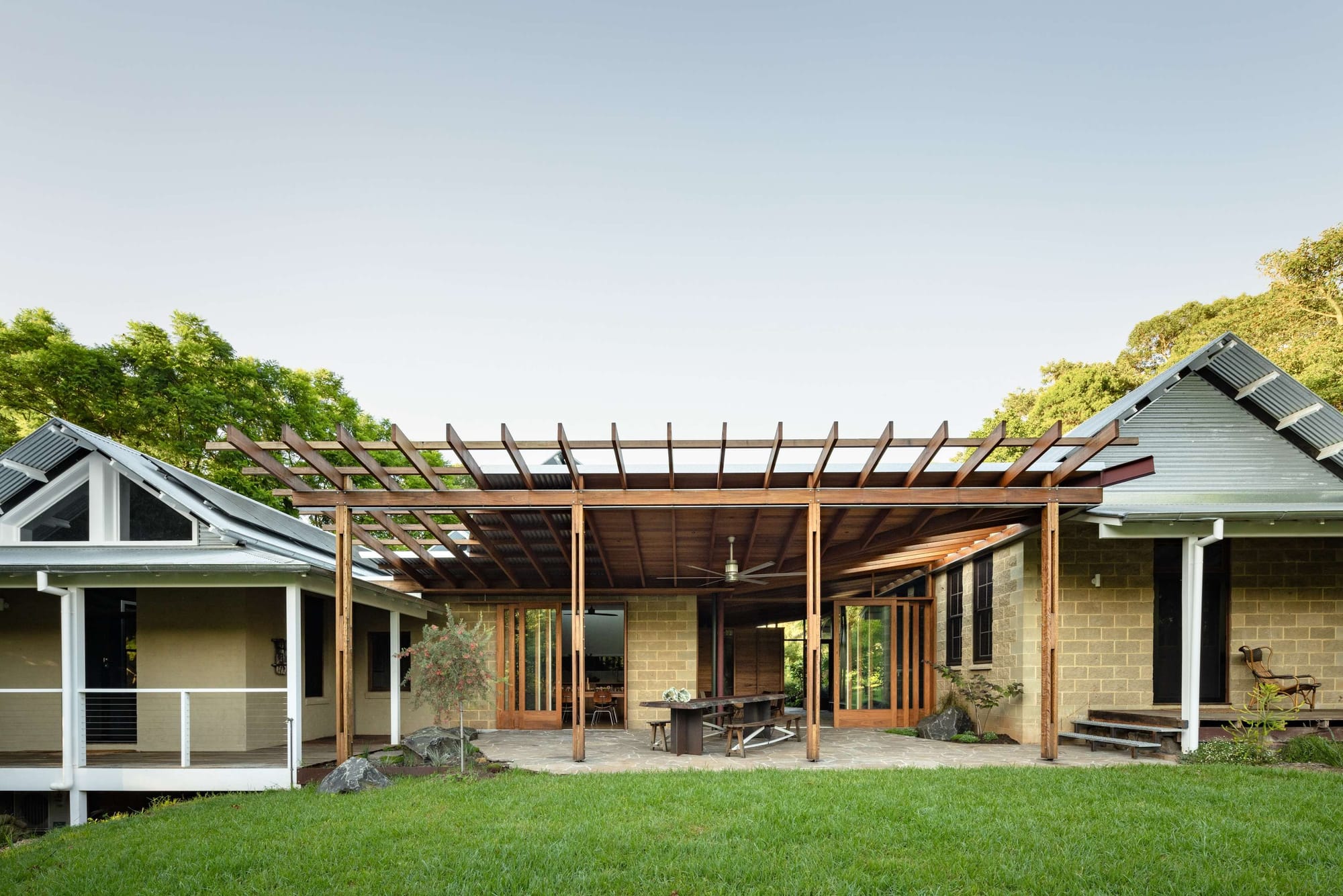 A broad timber verandah connects gabled pavilions, sheltering an outdoor dining table beneath a ceiling fan. Stone paving meets a green lawn and established trees.