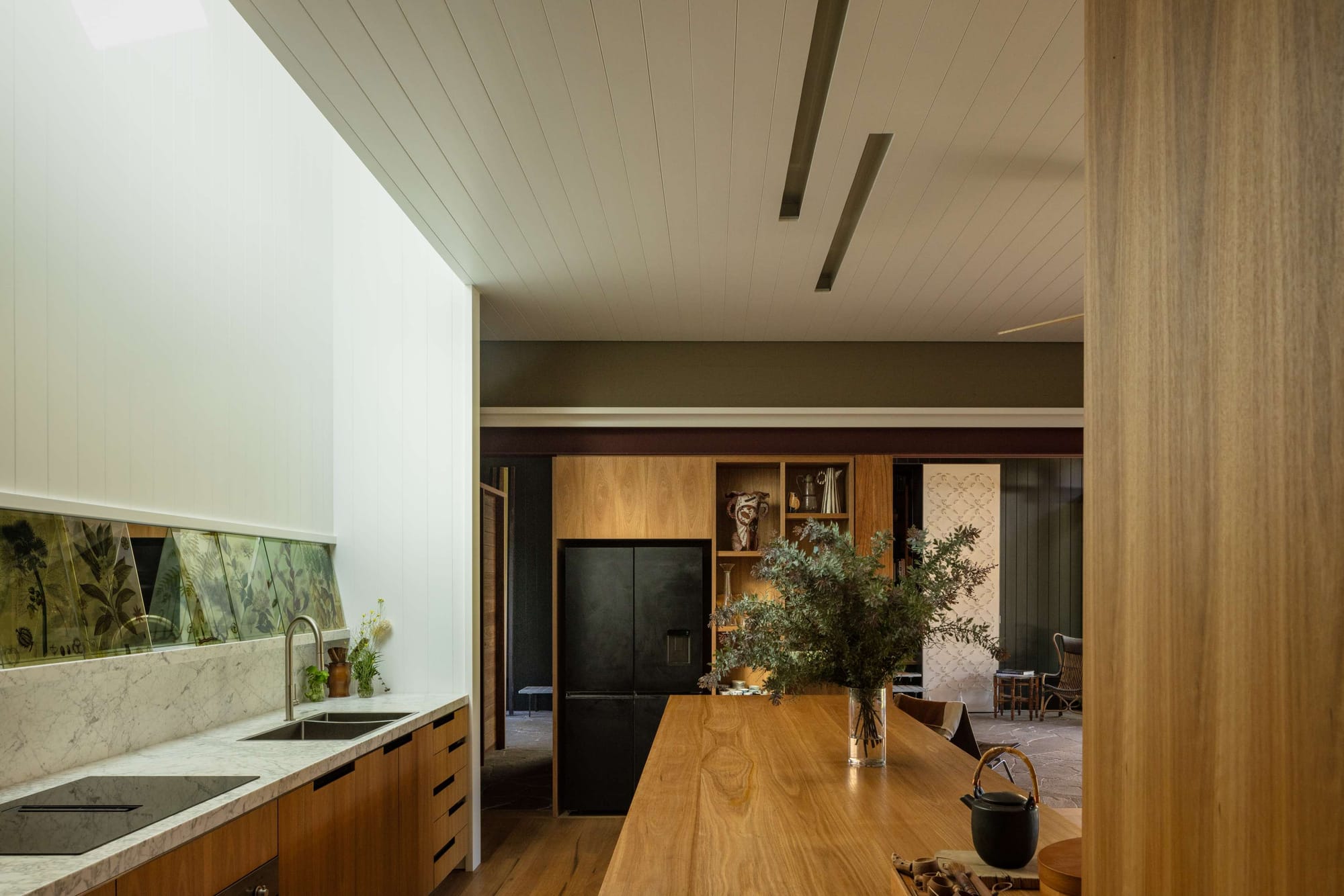 Natural light illuminates a kitchen with timber cabinetry, a long timber island and a pale stone benchtop. Botanical artwork runs along the splashback beneath a tall white wall.