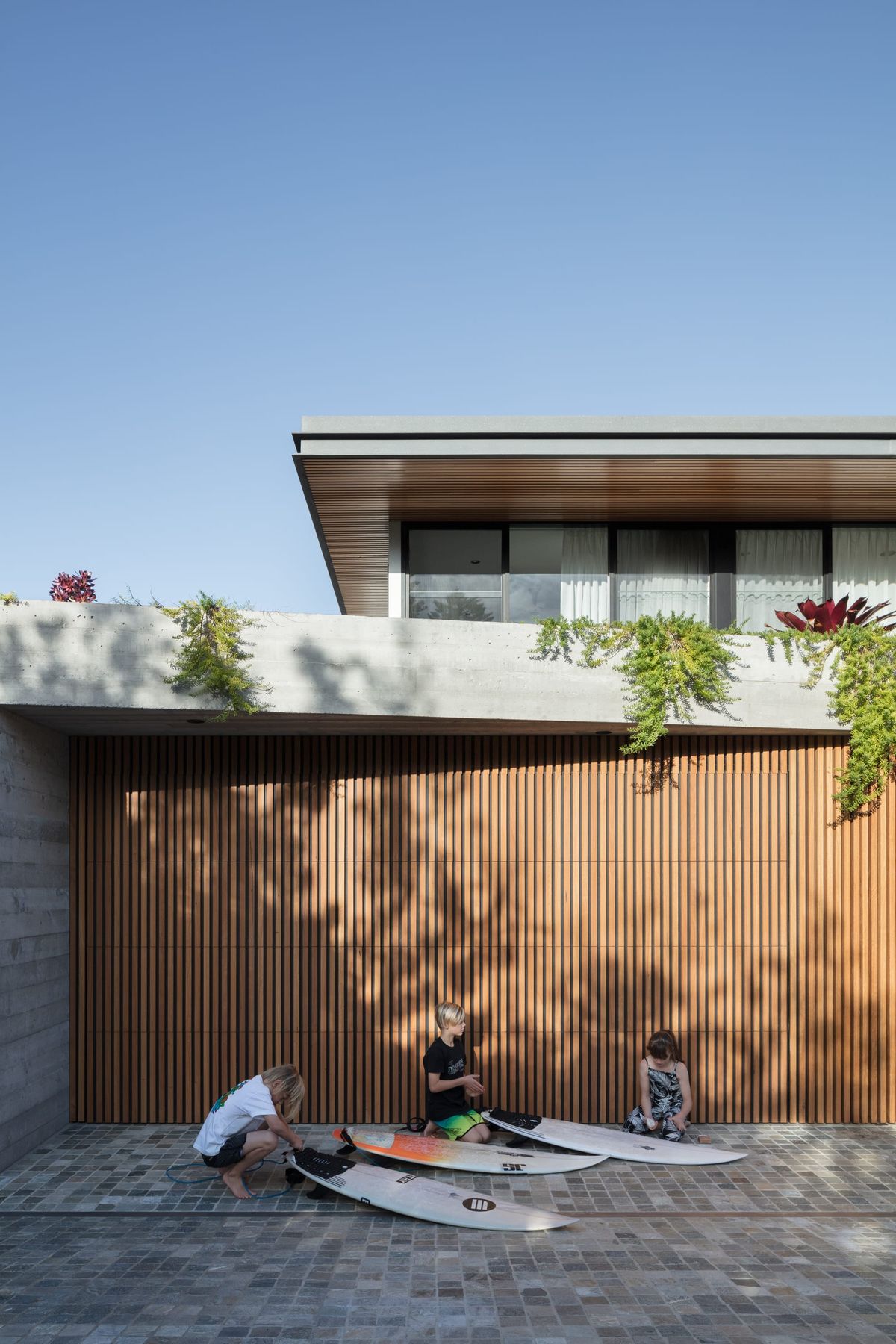 Showing the facade of the house with three kids waxing surfboards on the driveway