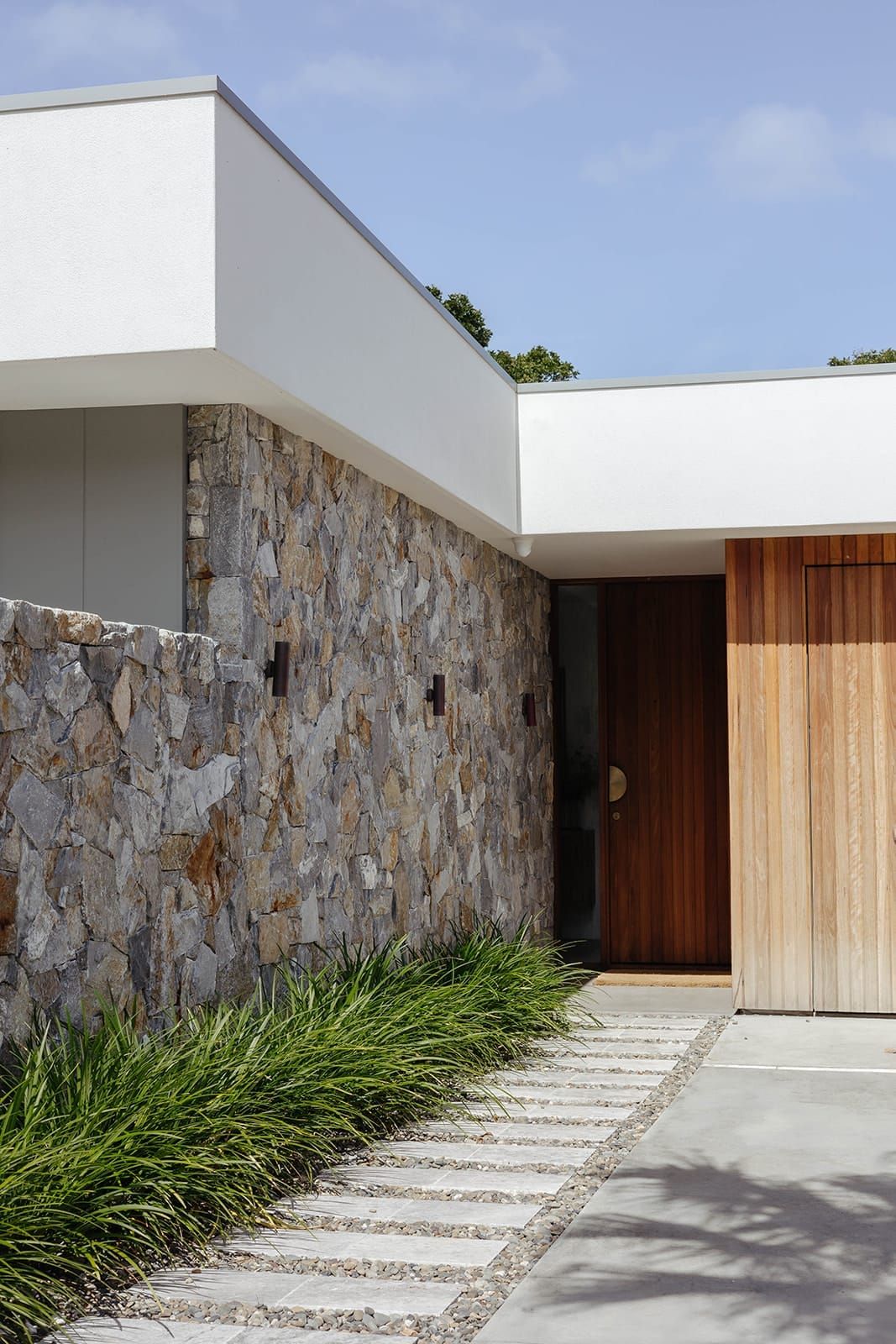 Street facade of home with timber door, stone wall and tiled footpath leading to entrance. Concrete driveway. 