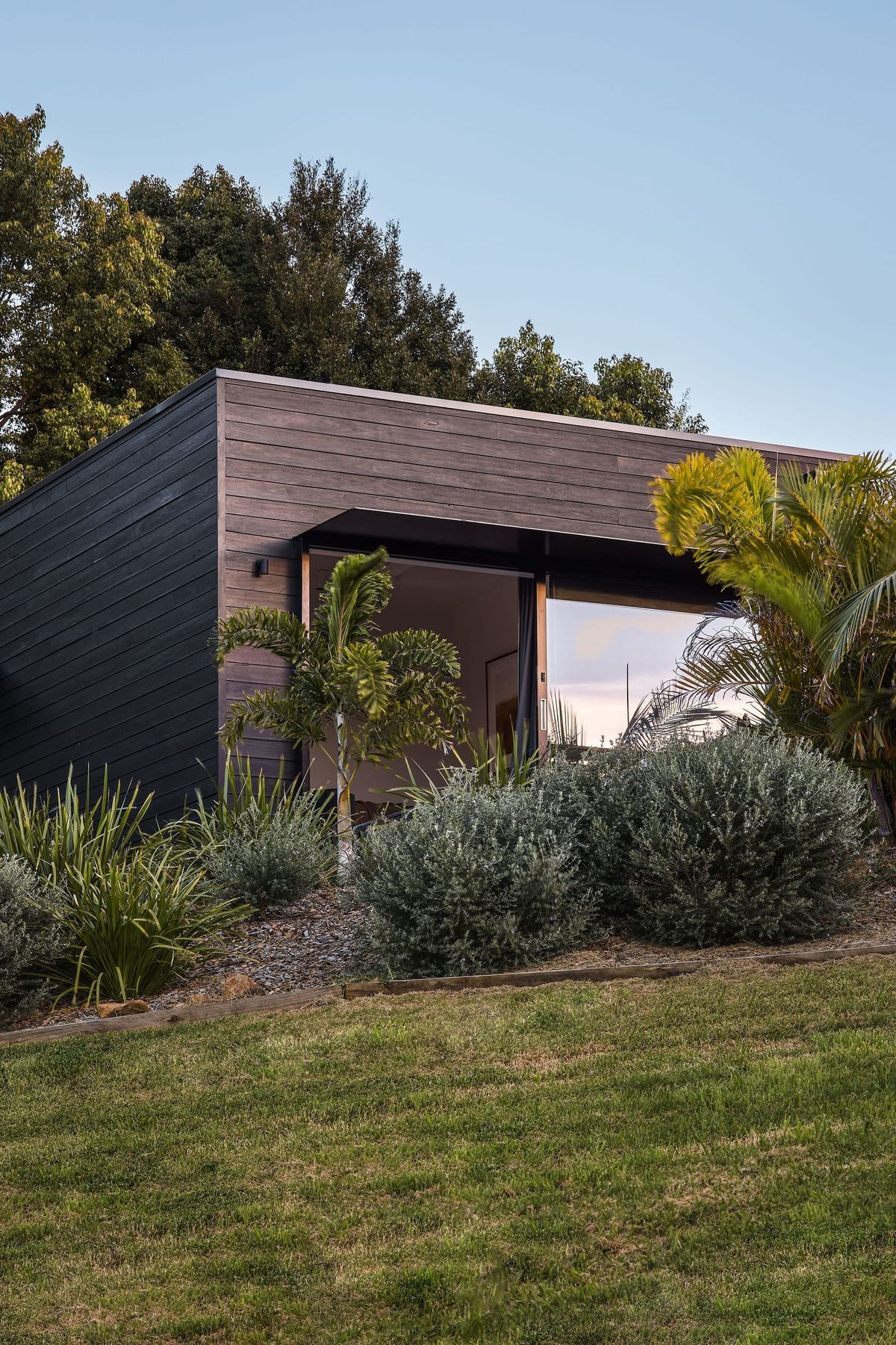 Square timber clad structure with large open windows. Garden bed with array of trees and bushes in front of building