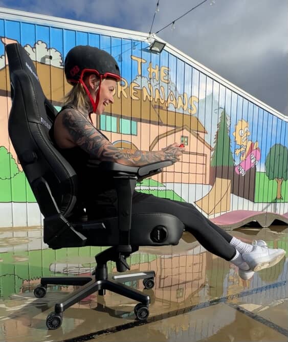 Retro Maggie gaming with a helmet on in a computer chair. There is a Simpsons mural behind her