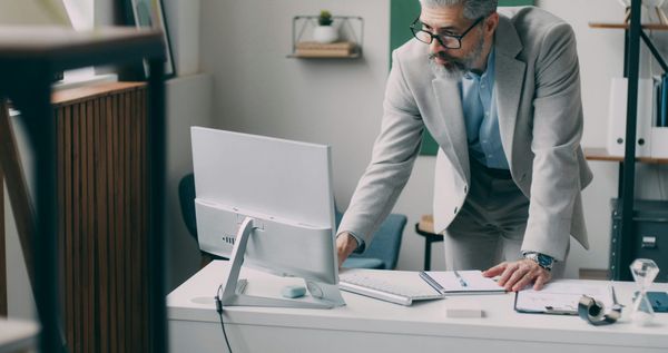Hombre de saco gris revisando la pantalla de una computadora, inclinado sobre un escritorio con documentos y una libreta.