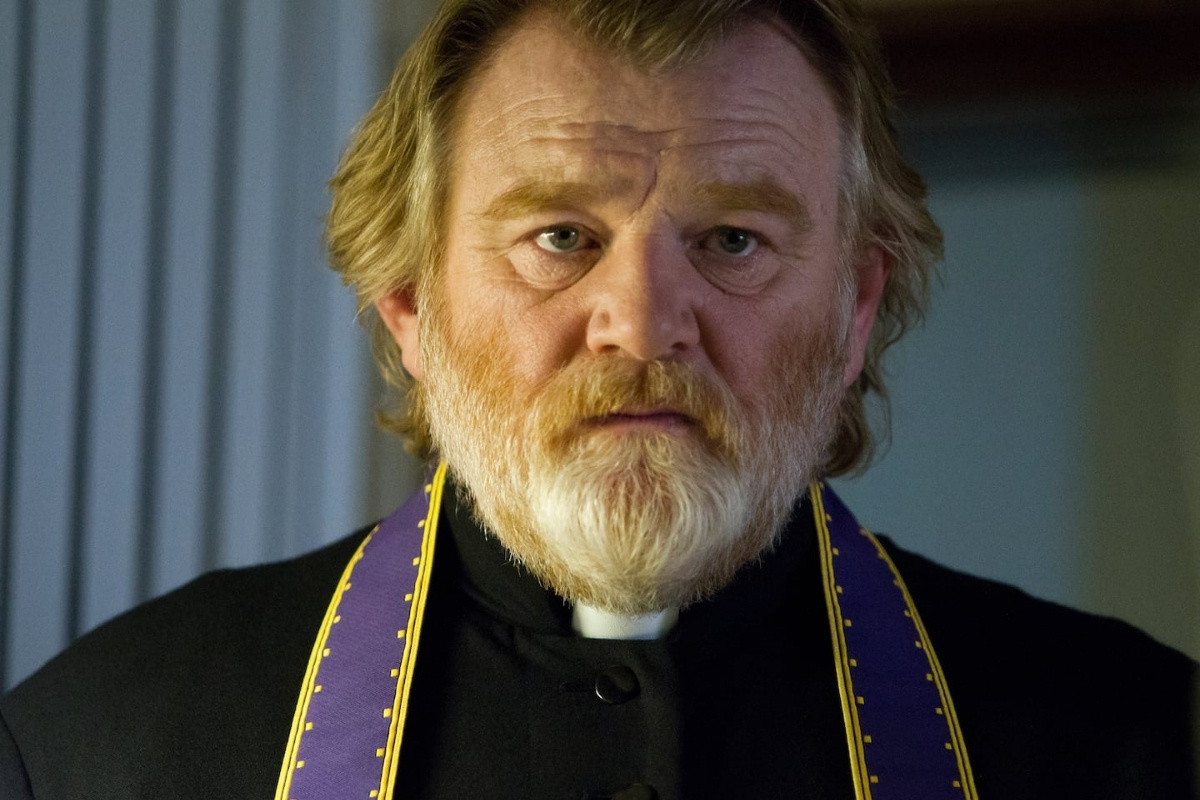 Brendan Gleeson as Father James in Calvary. He has a graying beard and wears a black clerical collar with a purple and gold stole, looking forward with a somber, weary expression.