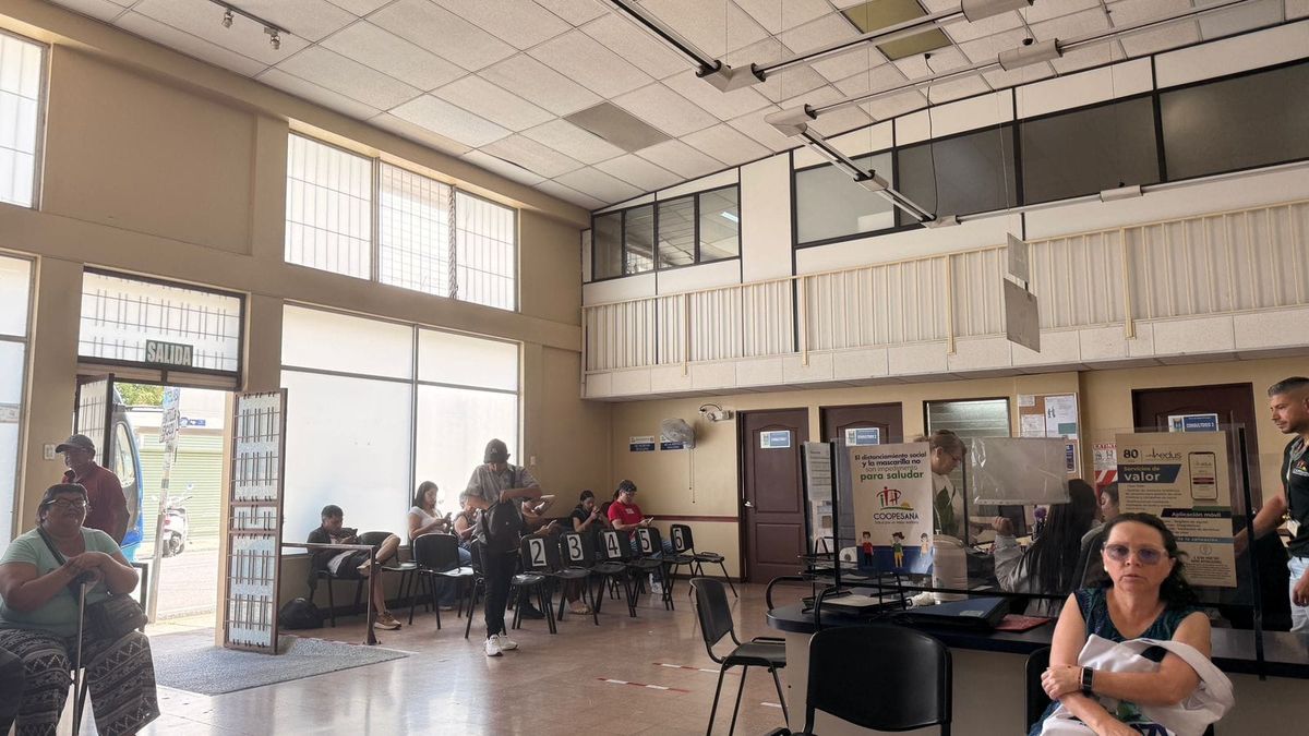 A sunlit waiting room in a Costa Rican EBAIS clinic. Patients sit in numbered black chairs under high ceilings. A plexiglass reception desk features COOPESANA and EDUS medical posters.