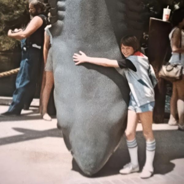 A smiling child in a denim vest and tube socks hugs a large shark statue at a theme park, likely the Jaws attraction at Universal Studios.