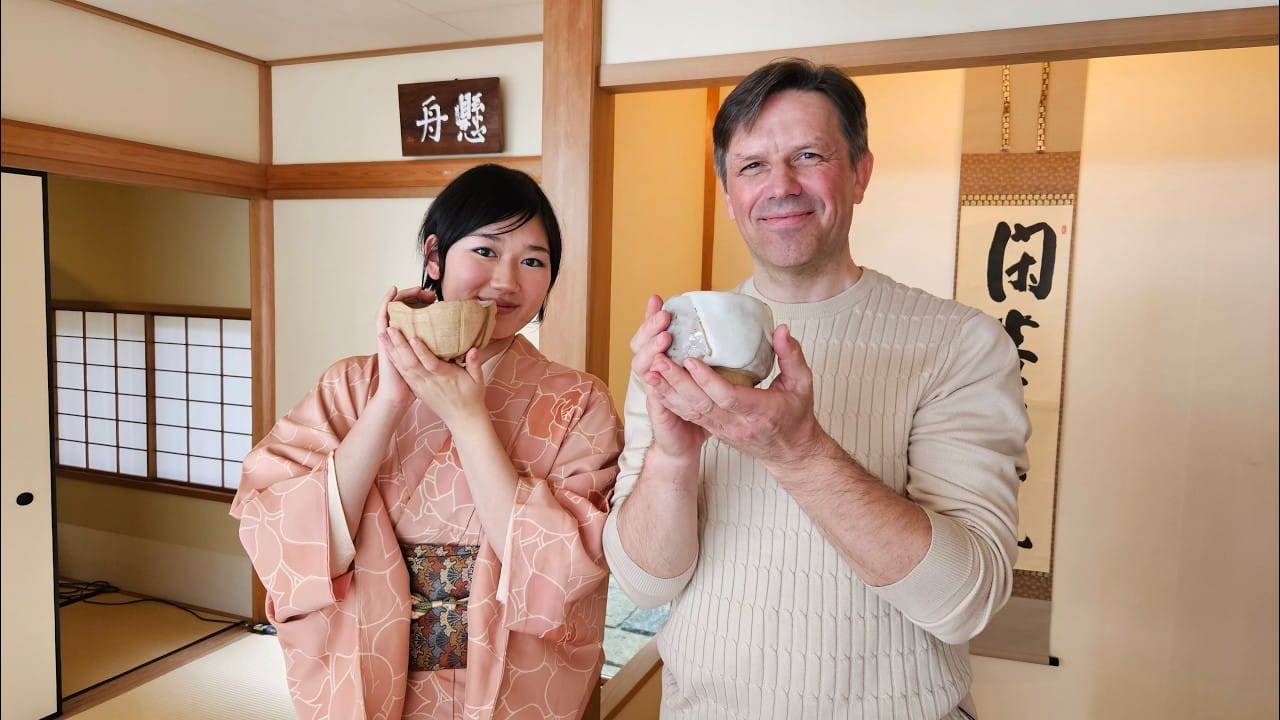Woman in pink kimono and man in cream sweater hold handmade tea bowls in traditional Japanese tea room with tatami mats and shoji screens