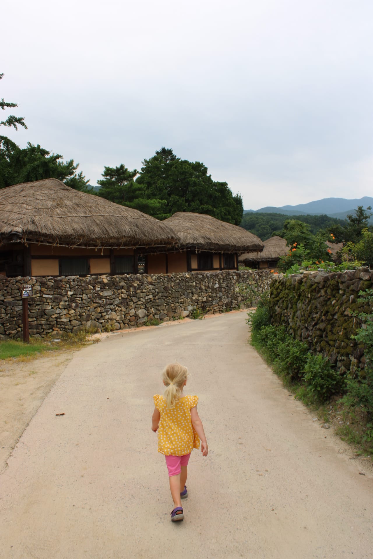 OeamFolkVillage_CreditAsheyWhitten_Street_girl Mädchen von hinten auf Dorfstraße zwischen Reetdachhäusern, Oeam Folk Village