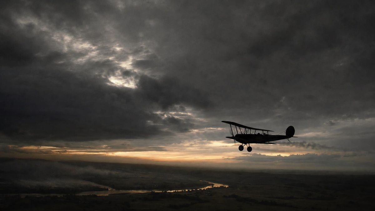 Vintage 1920s biplane in silhouette against a bruised twilight sky over rolling countryside.