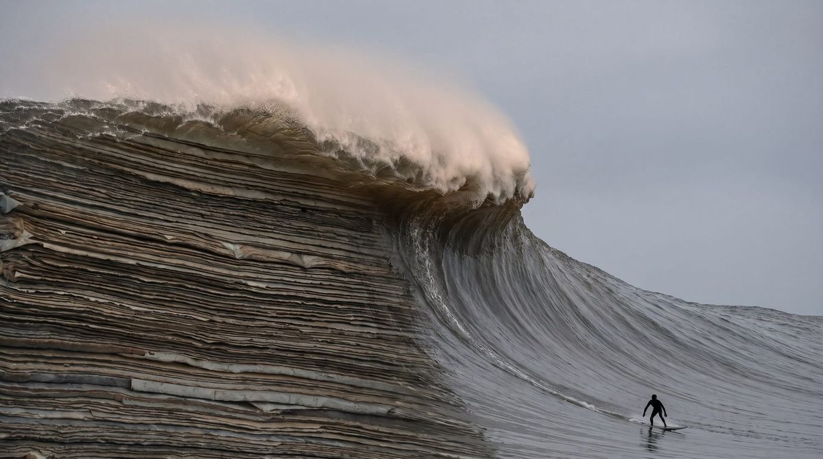 A monumental wave about to break, its face built of stacked strata resembling layered ledger paper, with a lone surfer far below on the smooth face ahead of the break.