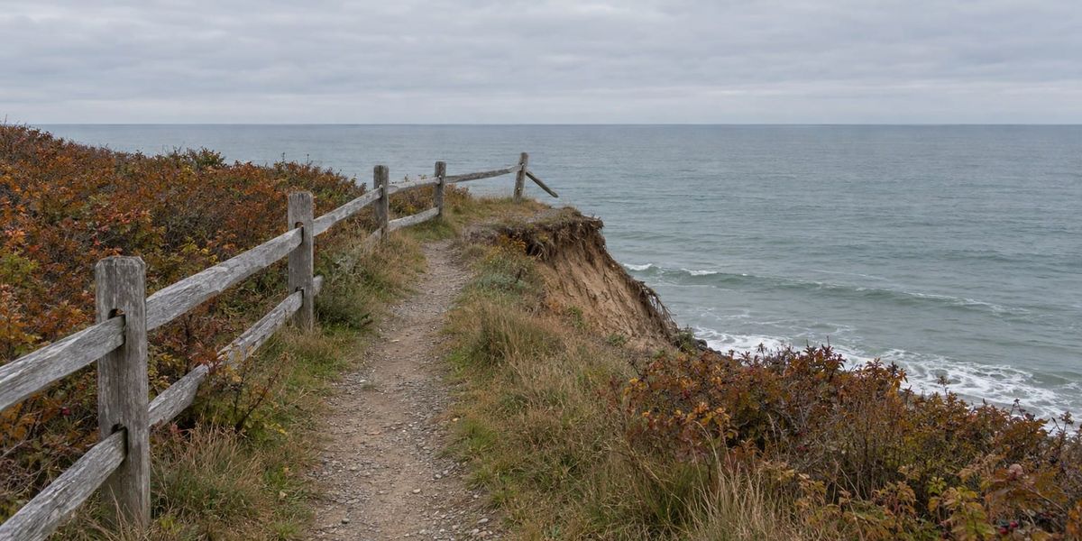 A gravel footpath along a coastal bluff on an overcast autumn day. The weathered split-rail fence beside it ends at the eroded edge, its last rail hanging loose above the sea.