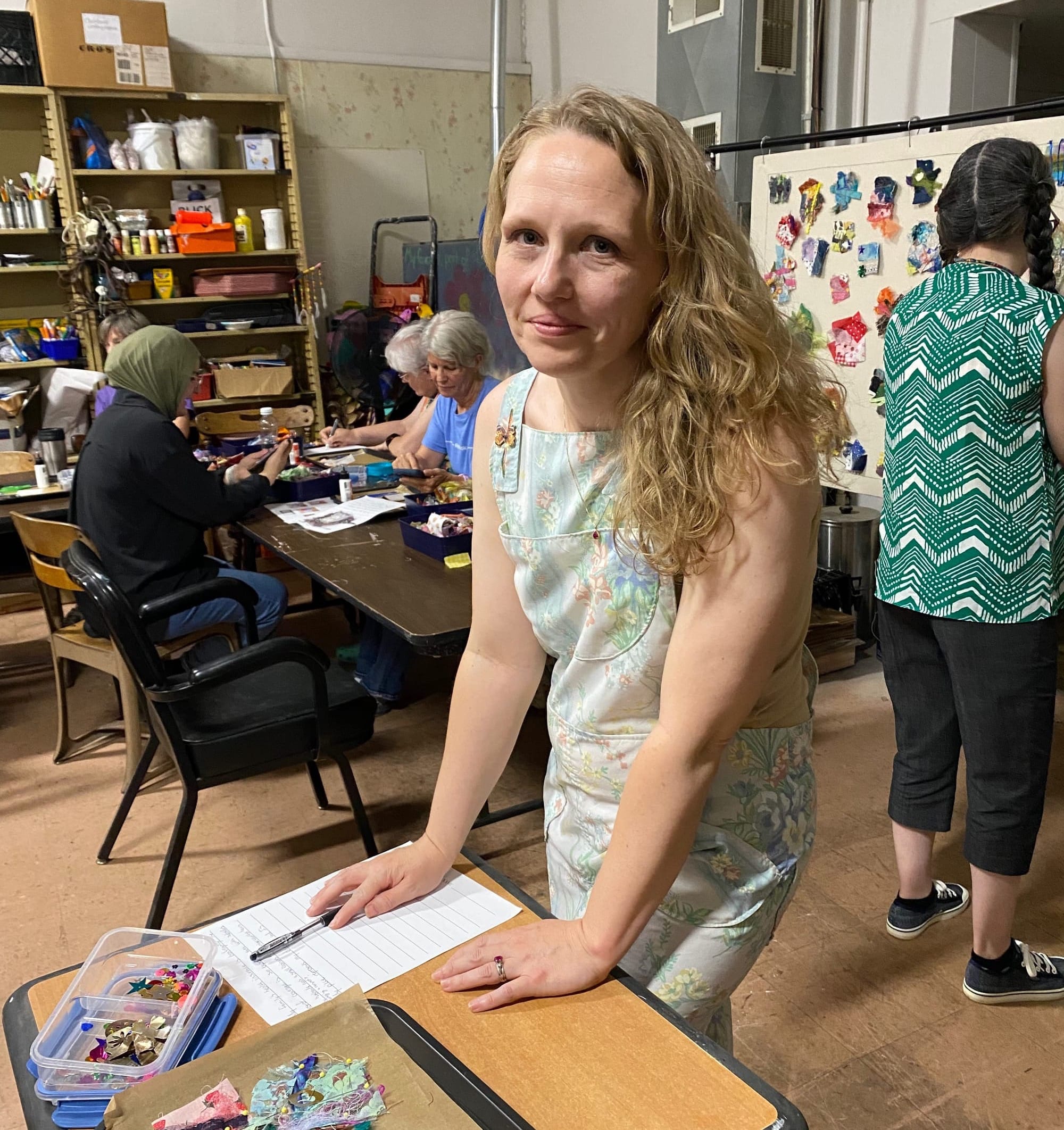 Woman grins and leans on a desk covered in art supplies. Behind her, other female-presenting people work on art proejcts.