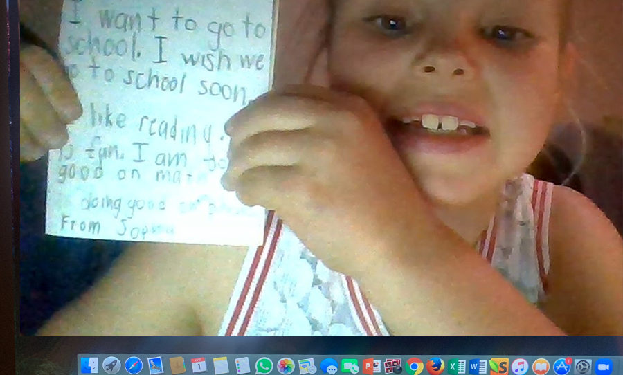 A young girl holds a letter up to the screen to let her teacher know she misses school.