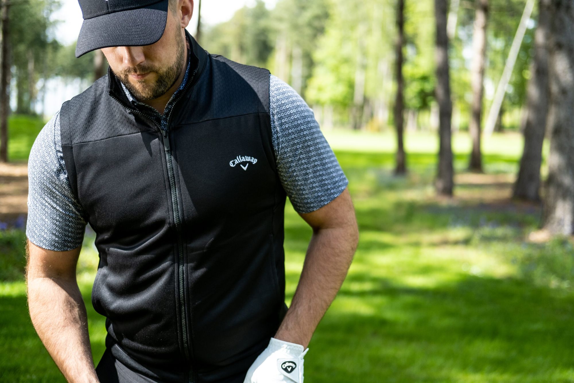 Male golfer wearing a gilet, polo, cap and golf glove with trees in the background