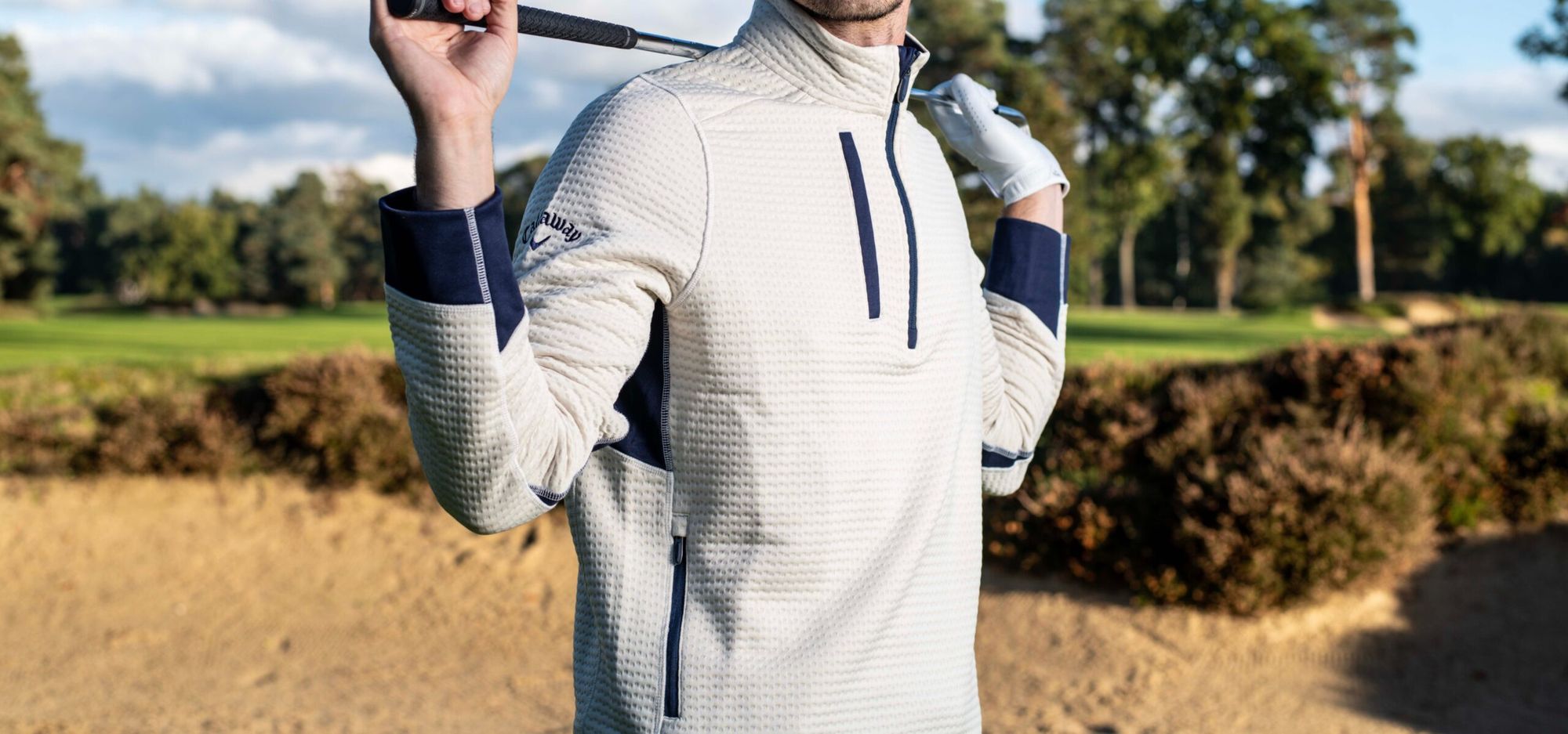 Male golfer standing in a bunker wearing a performance sweater with a club over his shoulders.