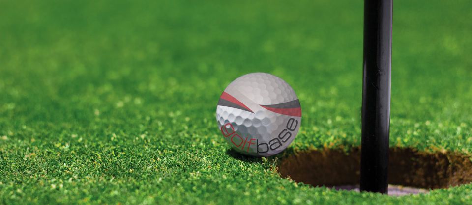 A close-up photograph of a golf ball with Golfbase branding on a golf green next to a hole with a flag post in it.