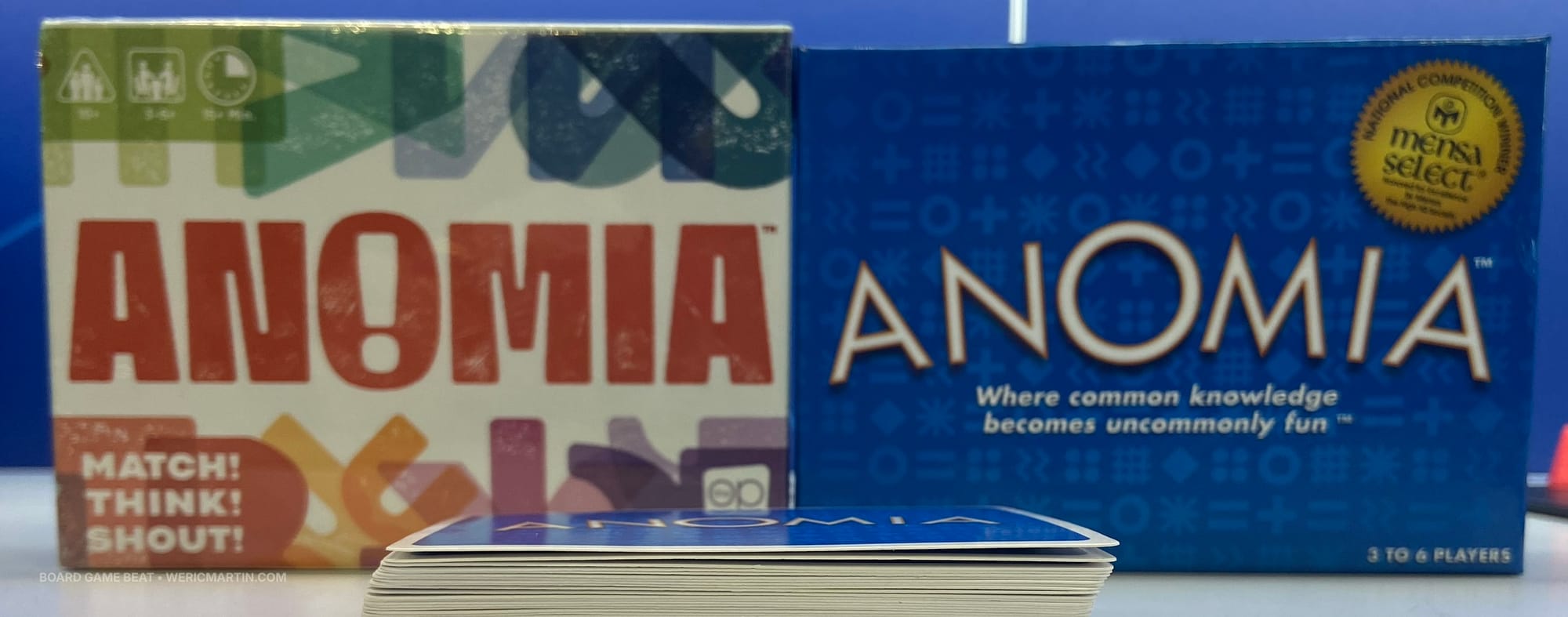 Two boxes of Anomia stand next to one another. The one on the right shows the older design with white letters on a blue box; the newer design at left is rainbow colored