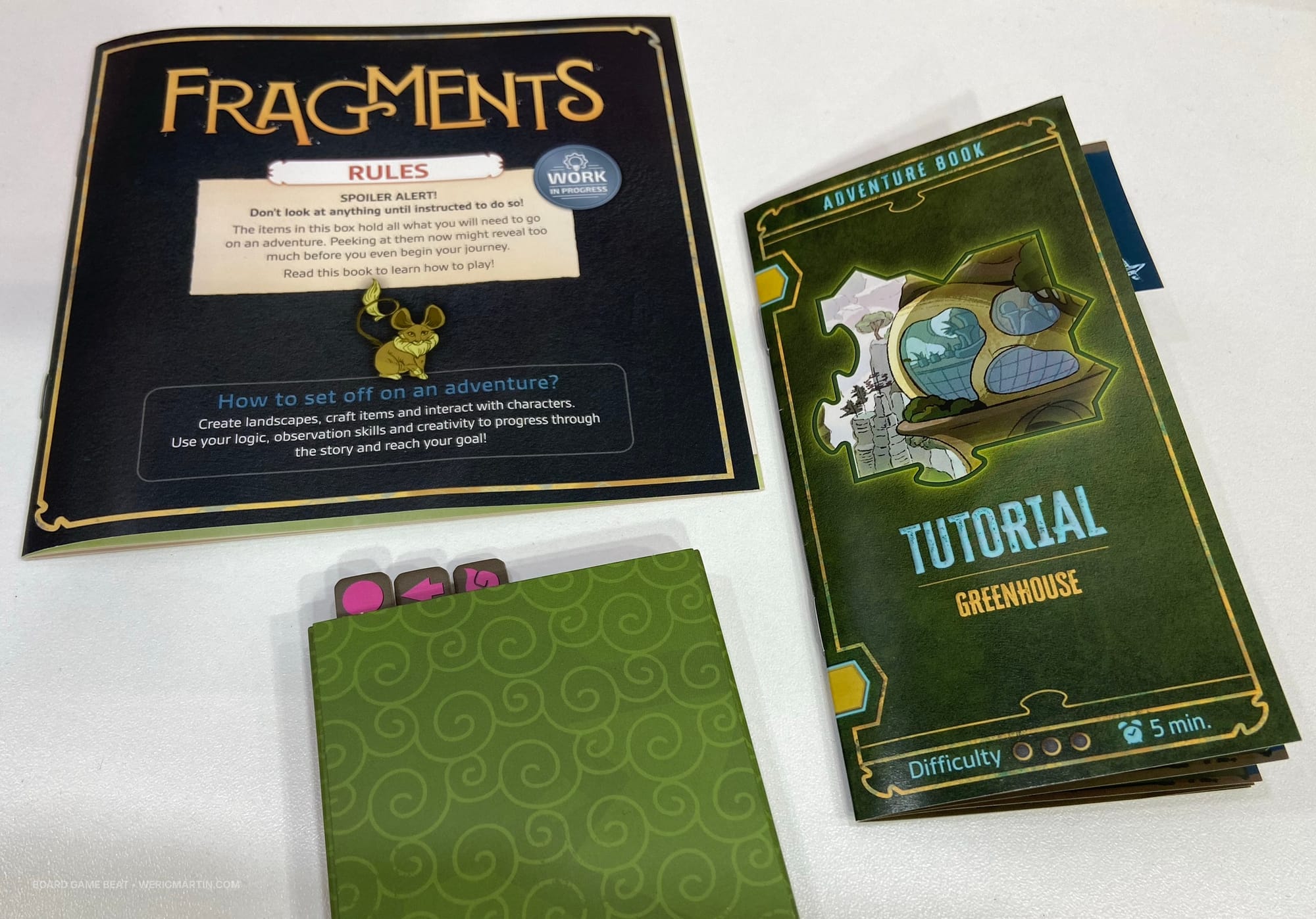A Fragments rulebook, adventure book, and cardboard sleeve containing three tiles