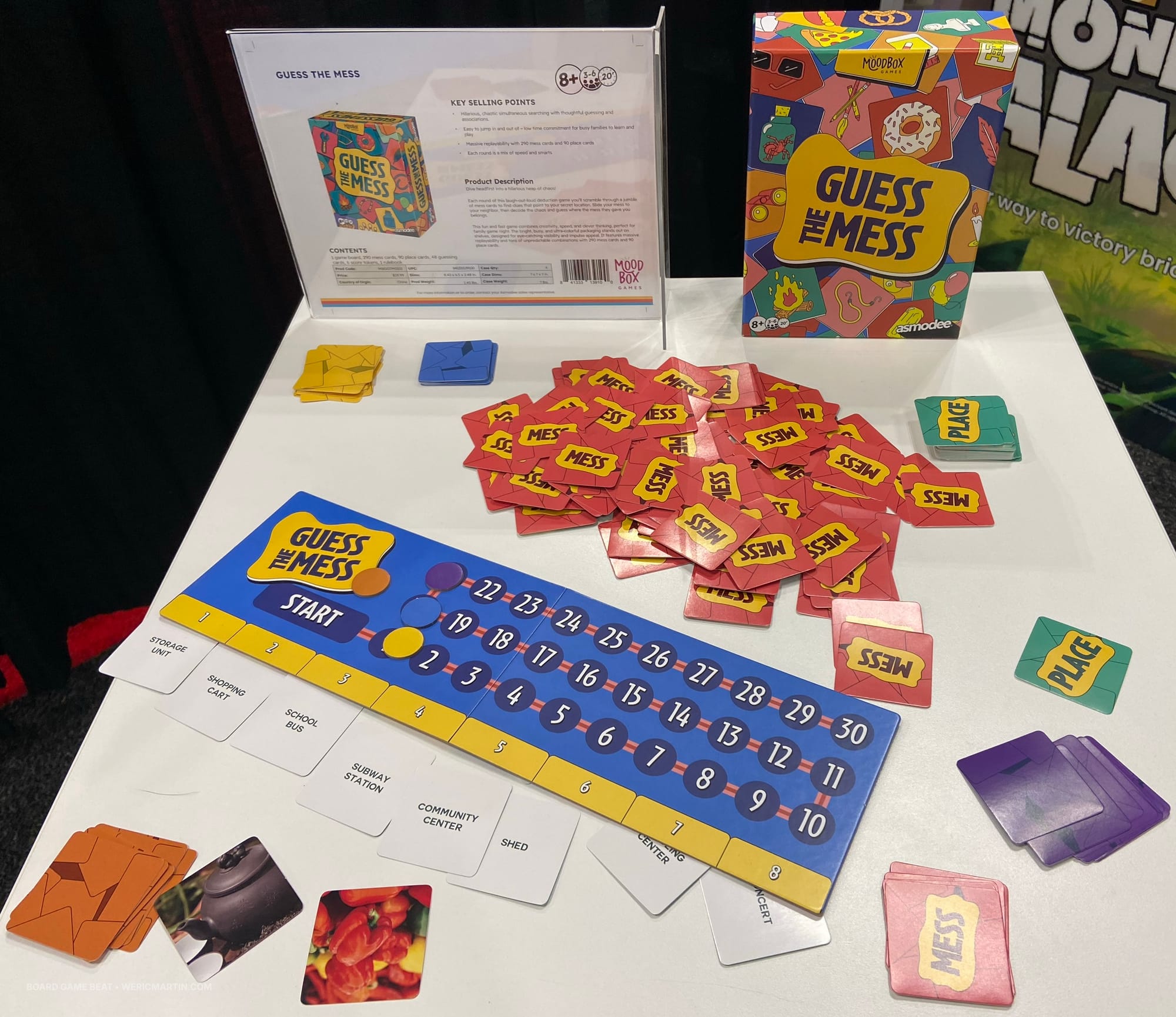 A messy pile of cards are in the center of the table next to the game box for Guess the Mess, a scoring track, and eight revealed "place" cards