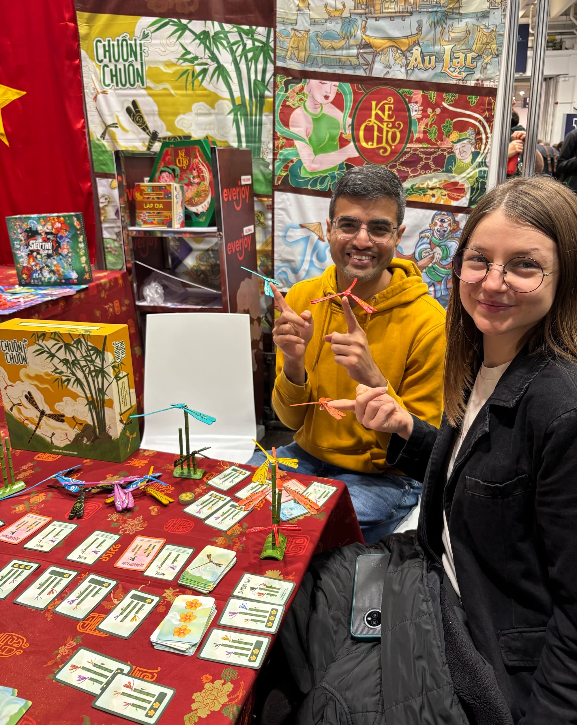 Two people balance bamboo dragonflies on their fingers, while sitting at a table covered with the components of Chuồn Chuồn