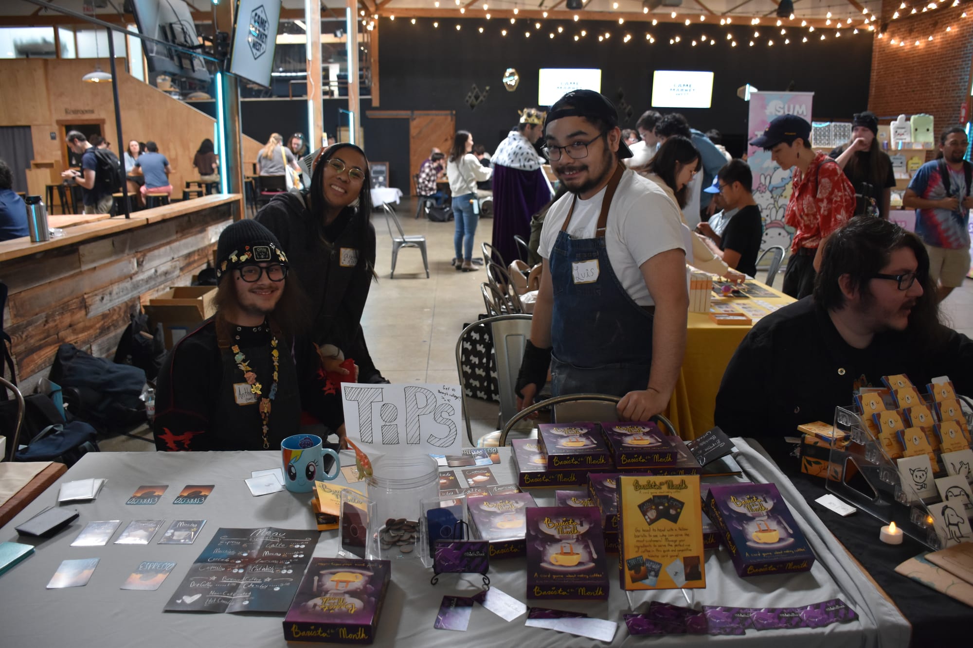 Designer Anthony Barbieri sits at a table bearing his game Barista of the Month, which is on the table available for a demo; two other people stand behind the table