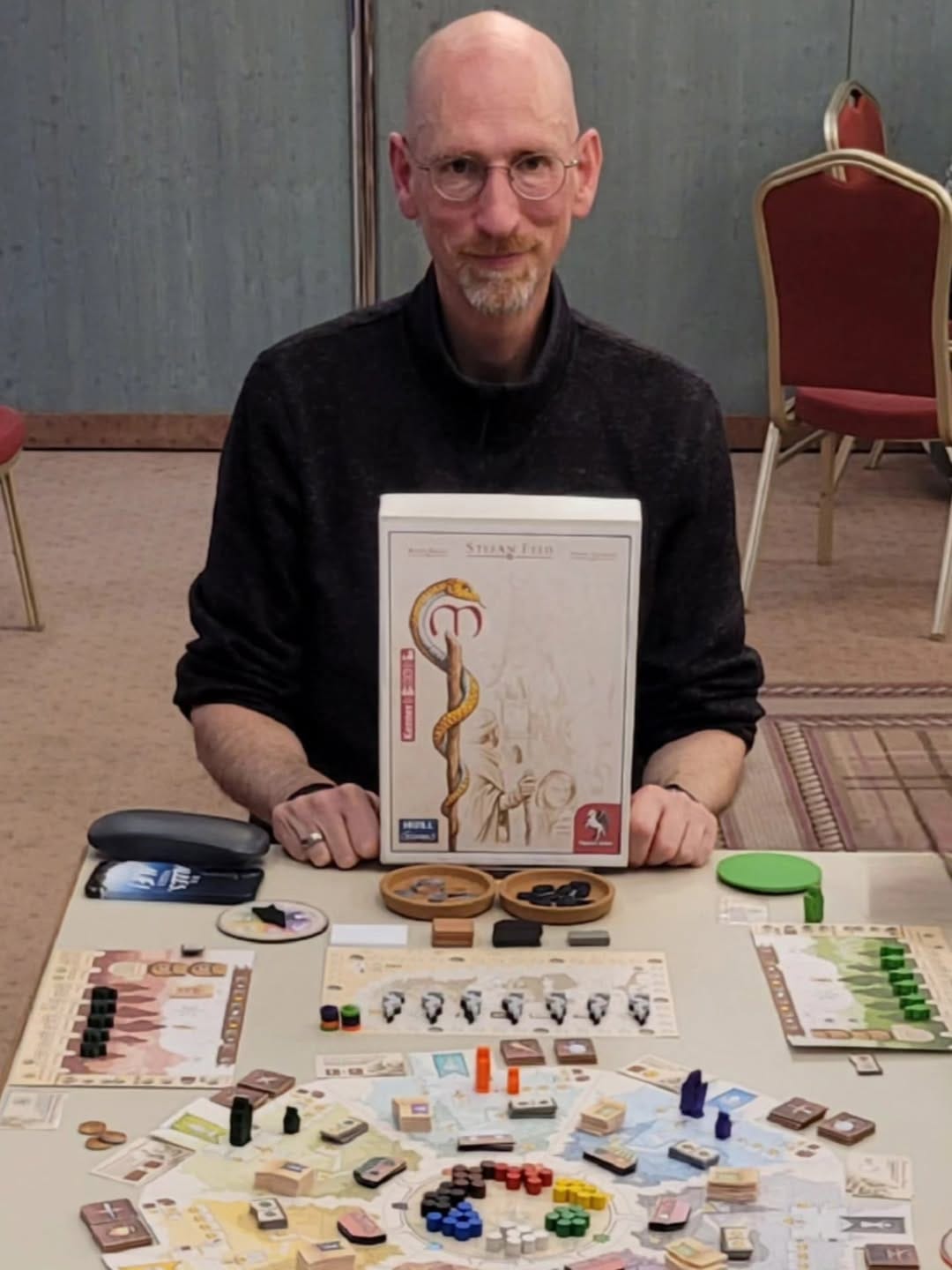Designer Stefan Feld holds a box top featuring unfinished art, with the table in front of him being filled with game boards, cardboard chits, and wooden components