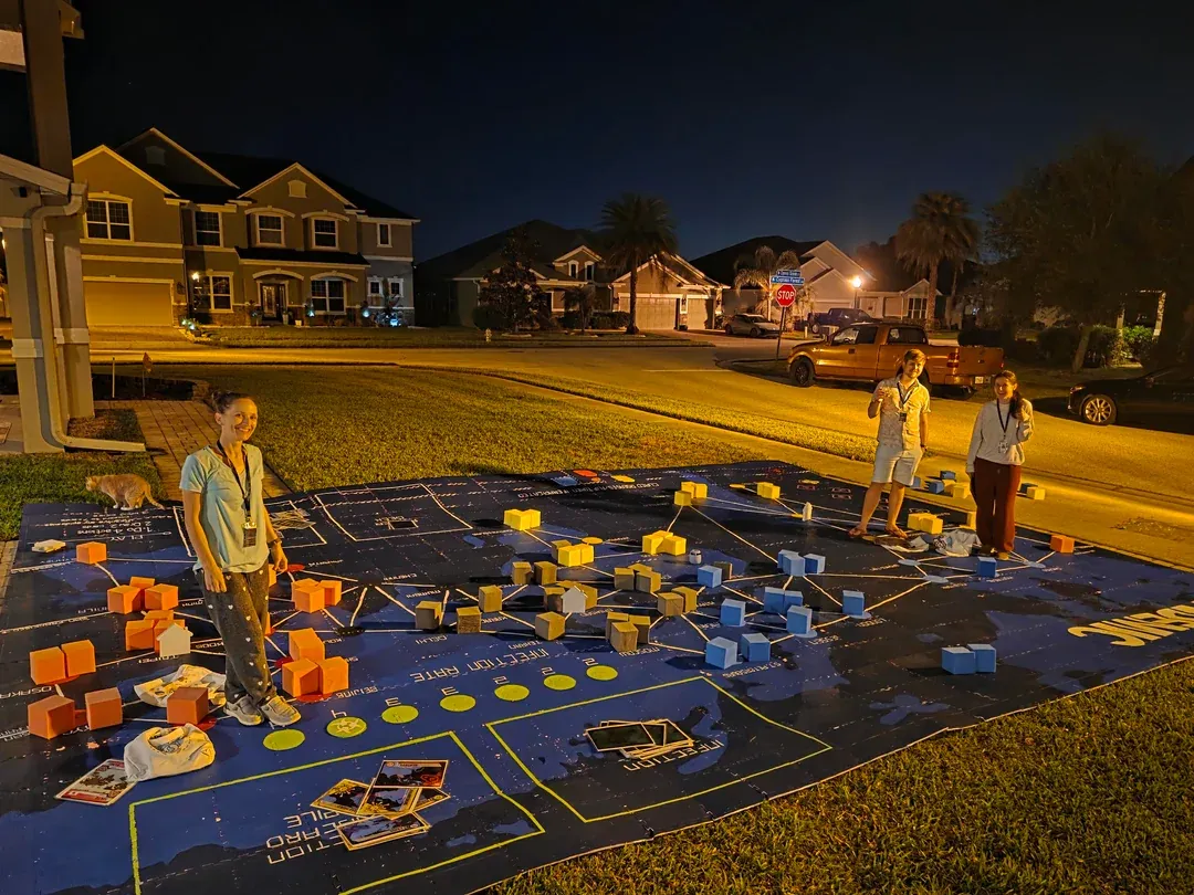 Three people stand on a Pandemic game board created from foam tiles that covers the driveway in front of someone's house