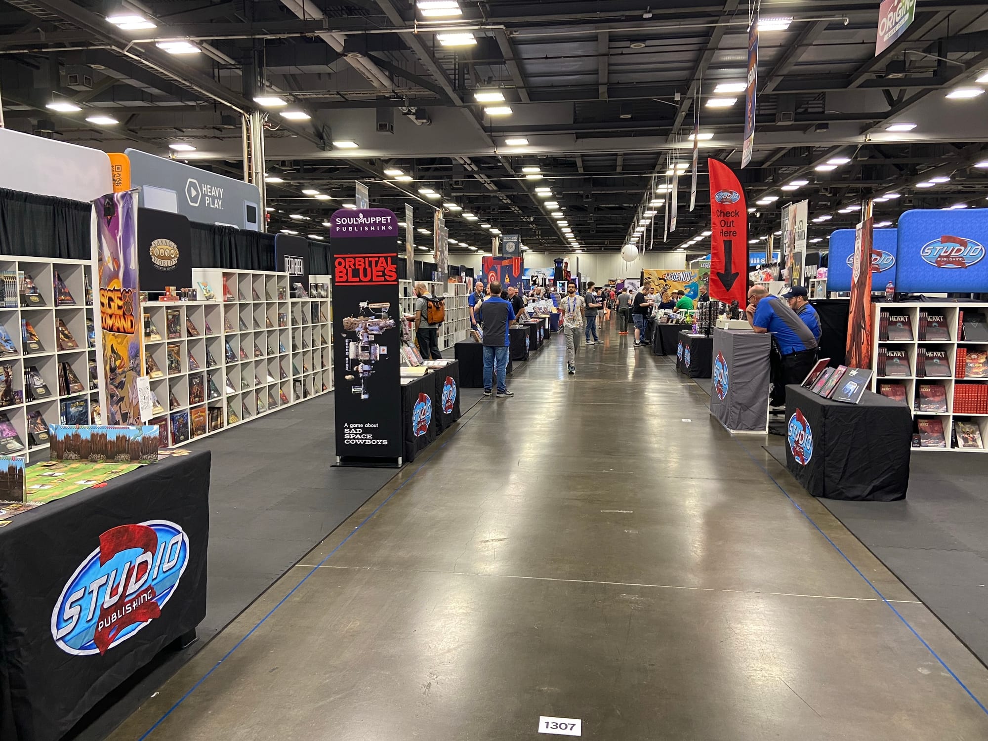 A view of a horizontal aisle at Origins Game Fair 2026, with a long row of bookshelves that block multiple vertical aisles