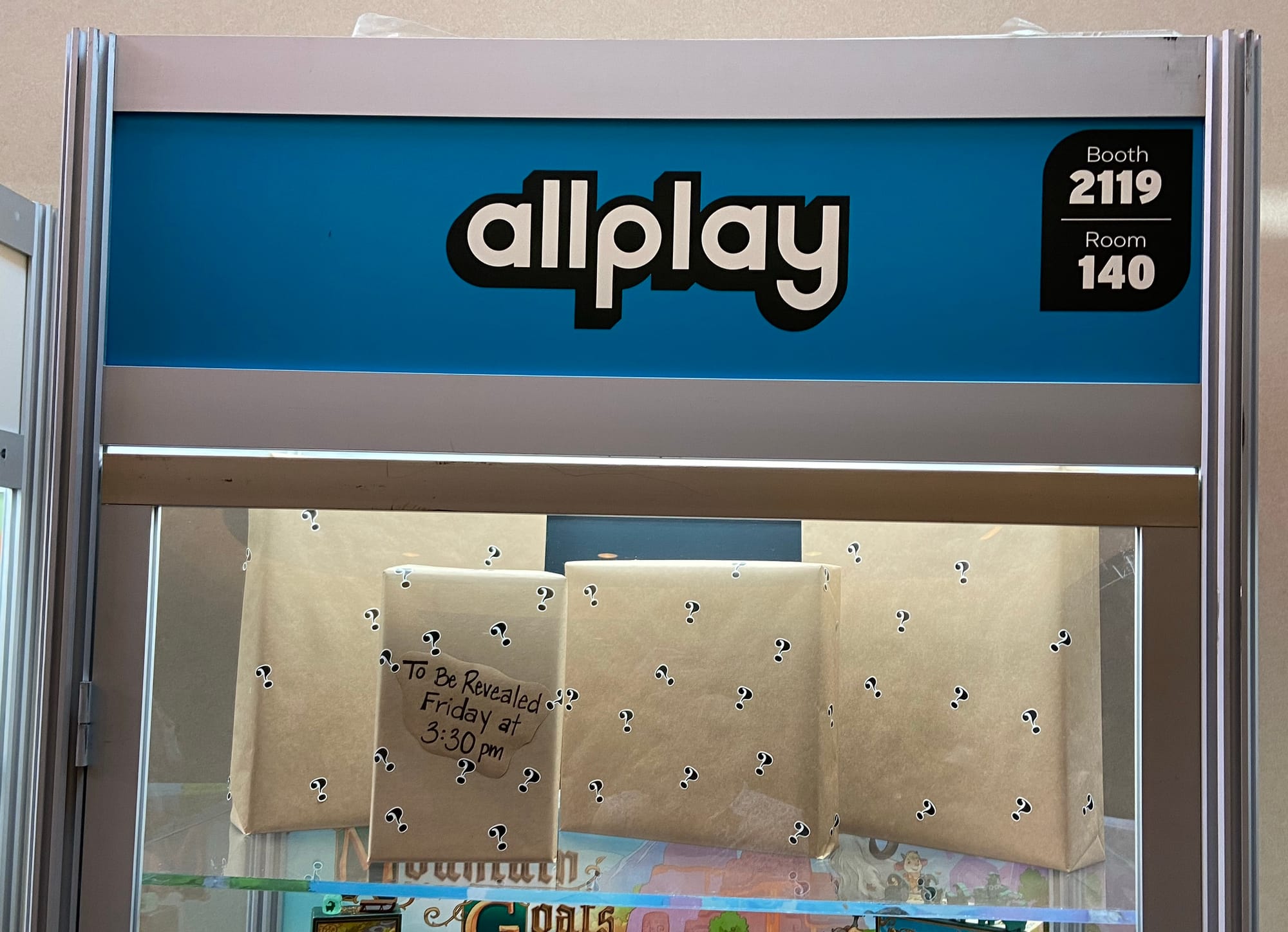 four games boxes wrapped in brown paper stand in a glass case with one bearing a sign that reads "To be revealed Friday at 3:30 pm"