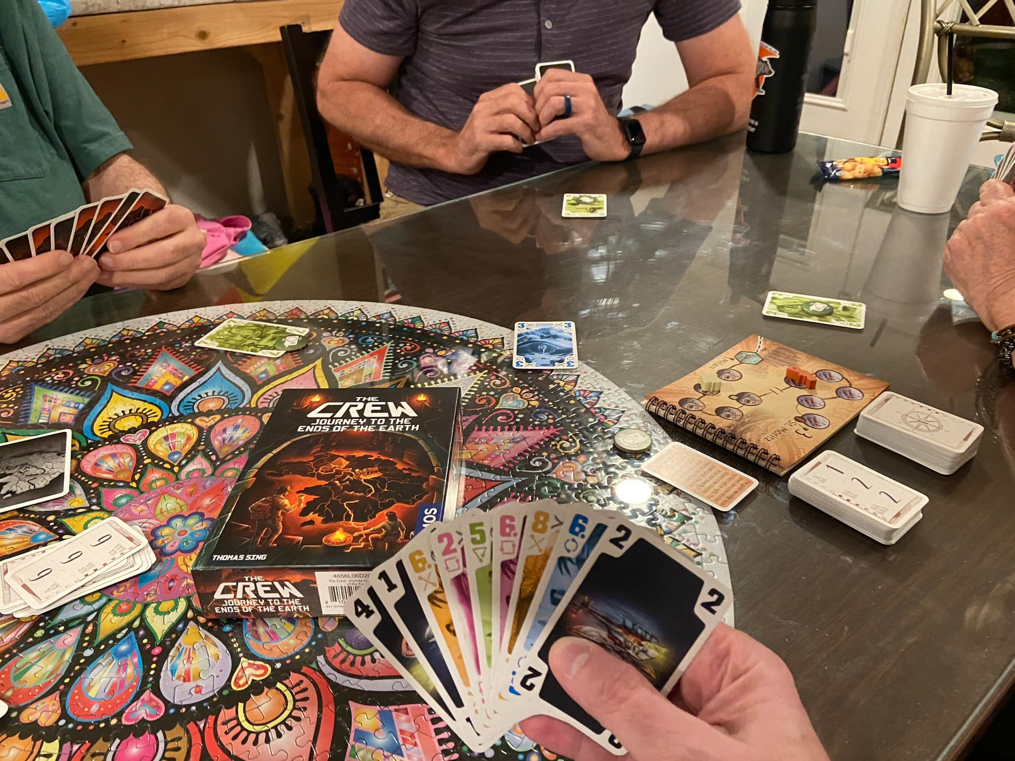 A hand of numbered cards in five suits is prominently placed in the image, in front of the box for The Crew: Journey of the Ends of the Earth; three other players are visible with cards of their own.