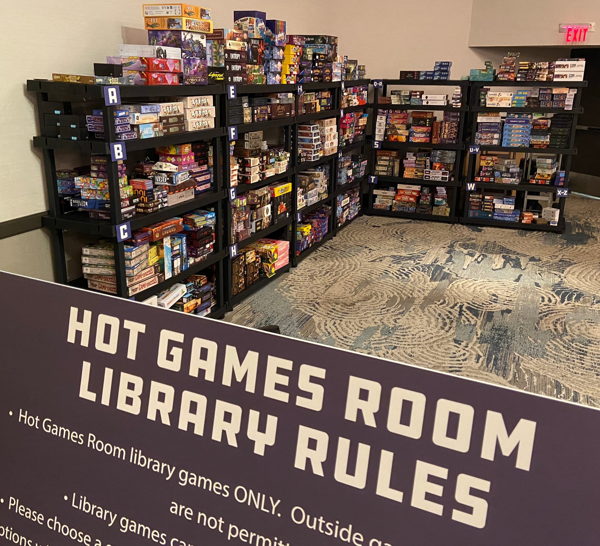 A sign reading "Hot Games Room Library Rules" is in front of six large plastic shelves filled with hundreds of games.