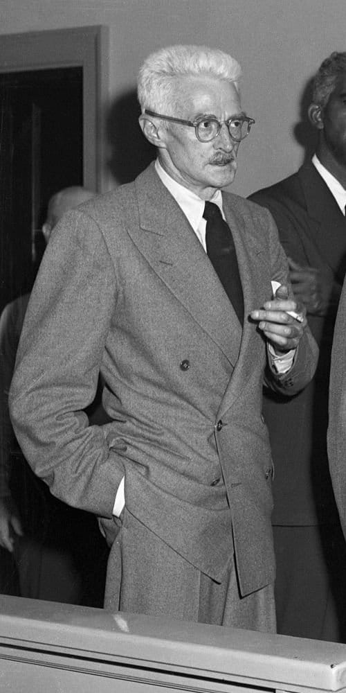 A man with white hair smokes a cigarette while posing in a suit with his other hand in his pocket.